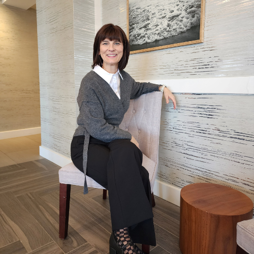 Woman sitting on a light-colored upholstered chair in a modern interior, smiling, with a wooden side table nearby and abstract wall art in the background.