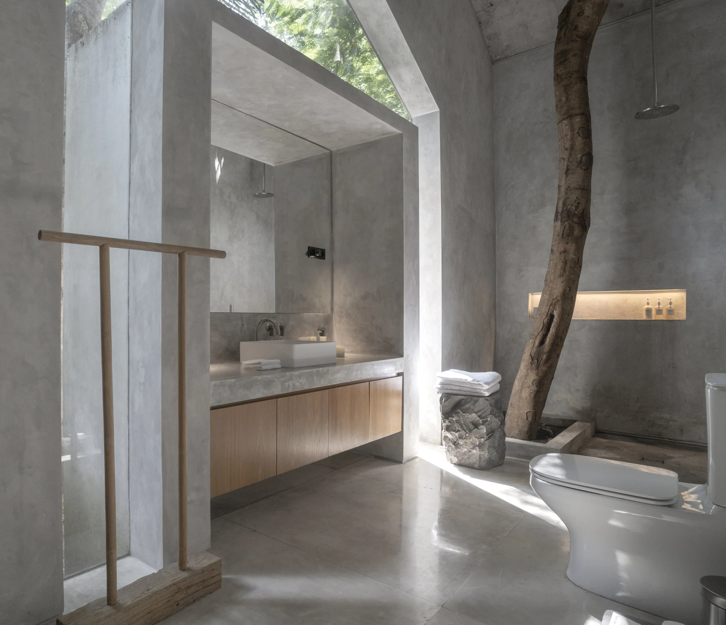 Unfolding Potential retreat. Modern bathroom with concrete walls and floors, a tree growing inside, a wooden towel rack, a stone with folded towels, a white toilet, and a washbasin with a mirror and recessed lighting.