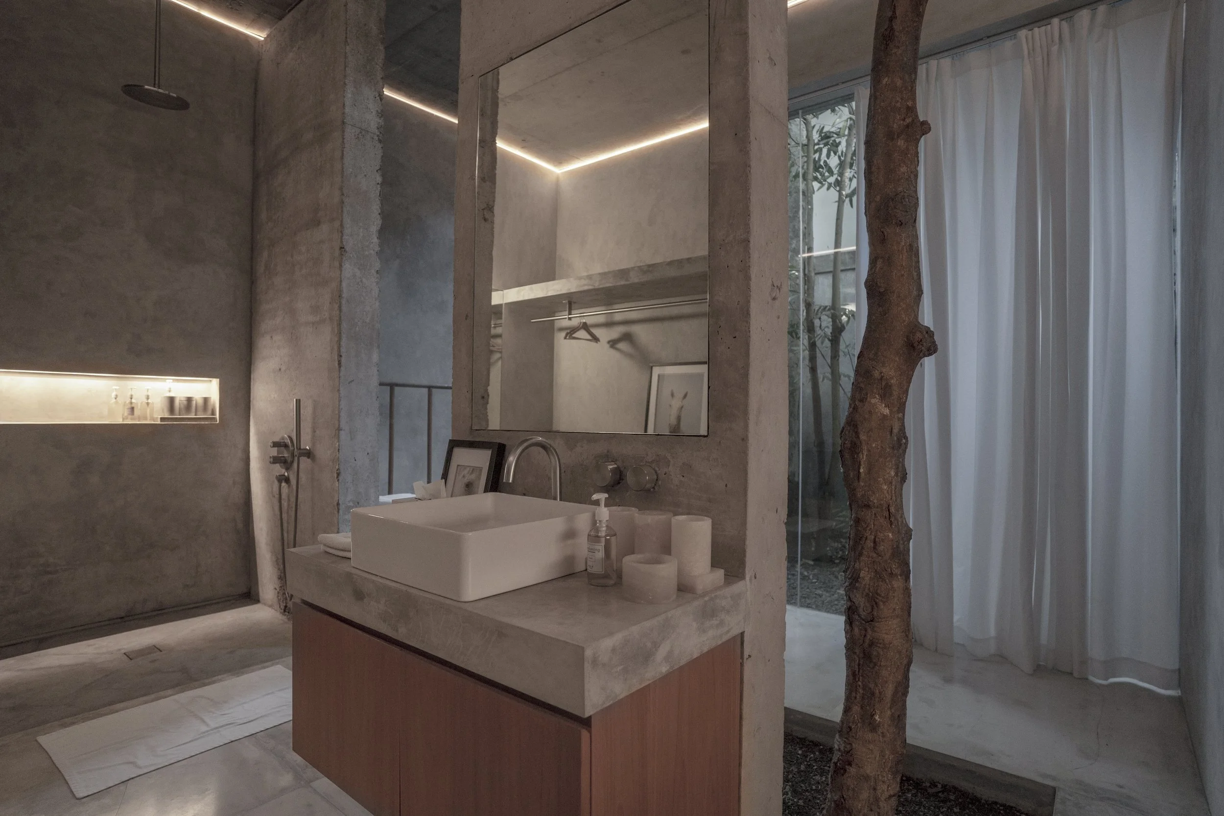 Modern minimalist bathroom with concrete walls, a concrete vanity with a white vessel sink, a large mirror, and a window with sheer white curtains. Features natural elements like a tree trunk near the window and subtle interior lighting.