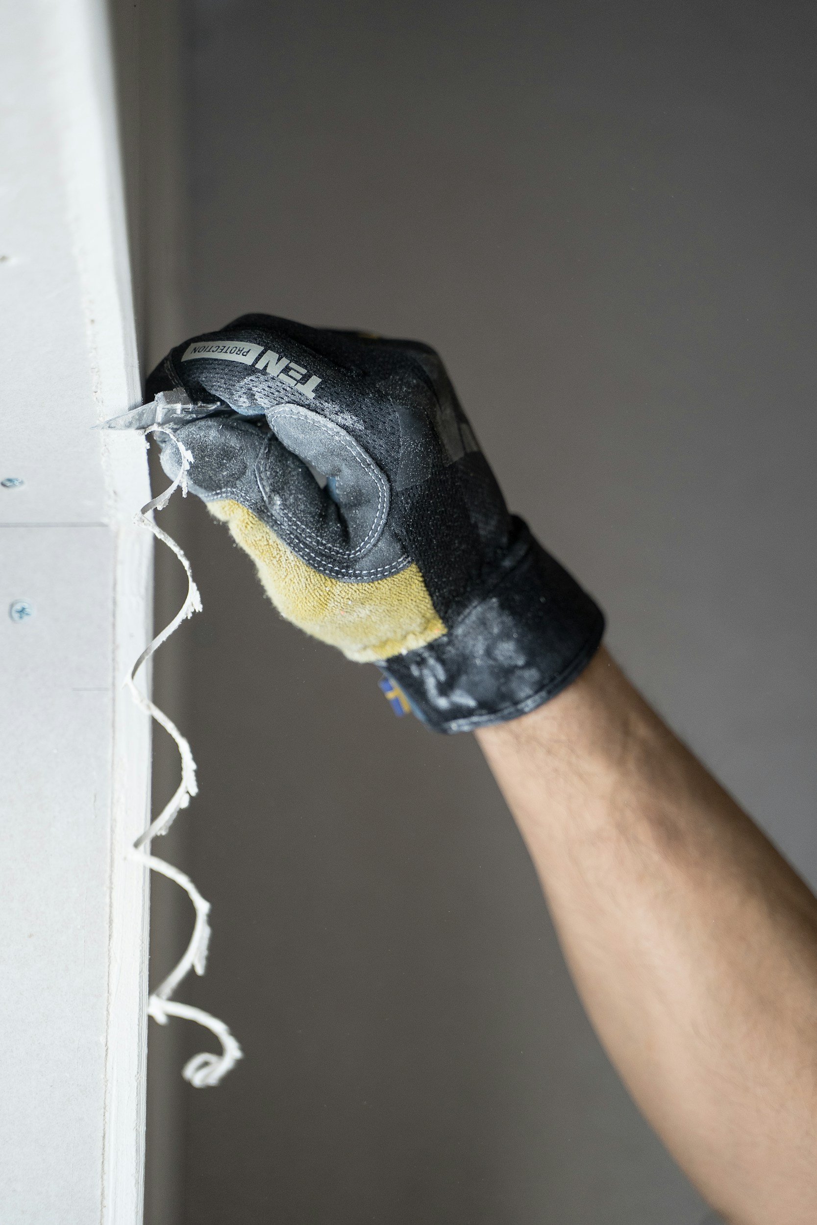 A person's hand wearing a work glove is installing or securing a piece of drywall to a wall or ceiling using screws. The hand is holding a screw, and the drywall has some white joint compound or primer on its edge.