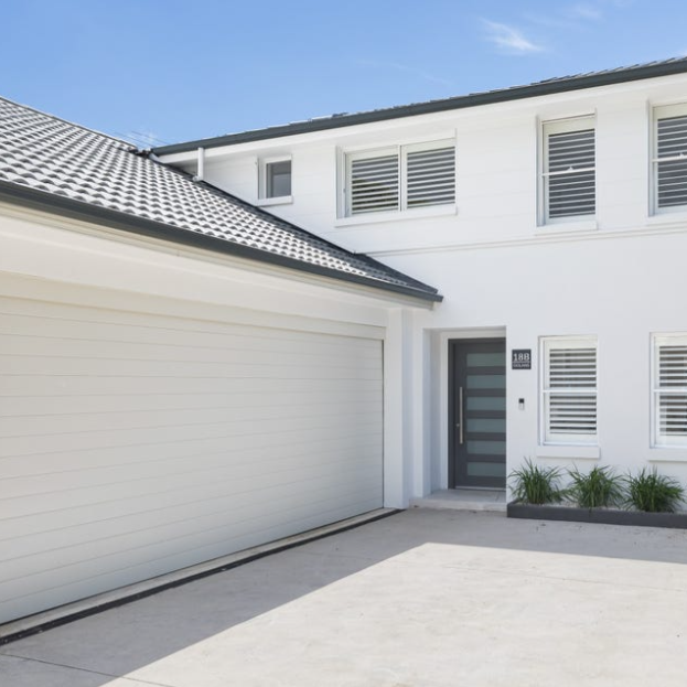 Modern white house with a closed garage door and a front door with horizontal glass panels, some small plants near the entrance, and a clear blue sky above.
