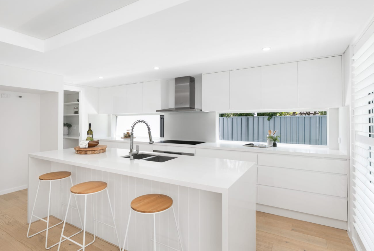 Modern white kitchen with island, three wooden bar stools, built-in cabinets, and a window with a view of a blue fence.