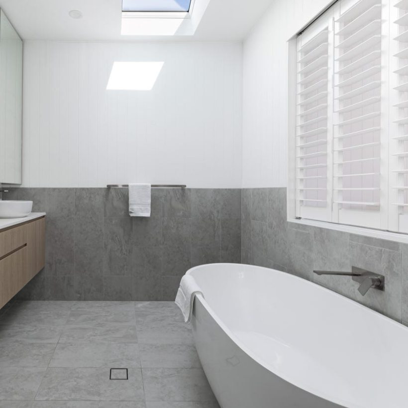 Modern bathroom with a freestanding bathtub, gray tiled walls and floor, a wooden vanity with a white sink, a towel rack with a white towel, and large white plantation shutters covering a window. Skylight provides natural light.
