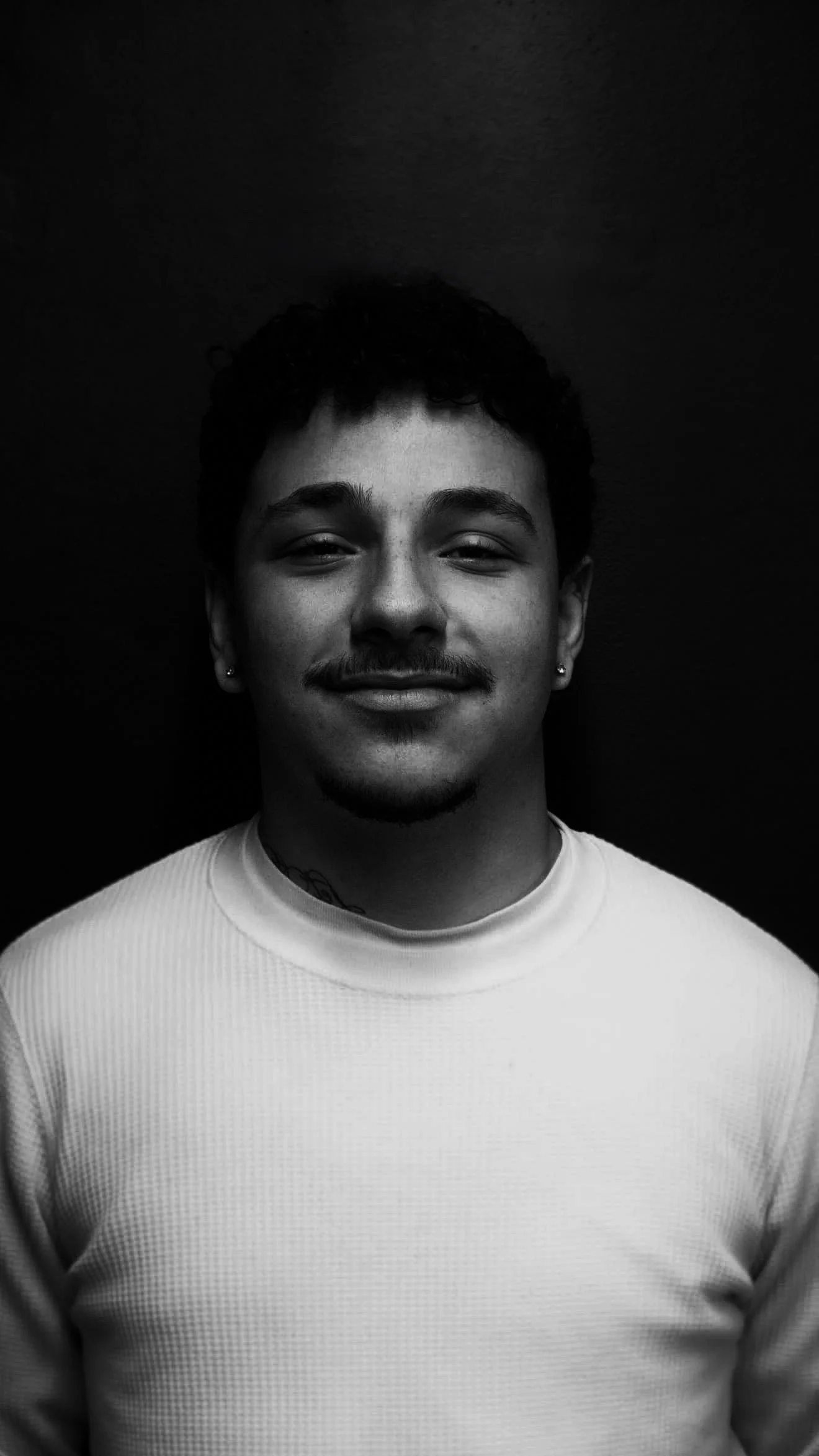 Portrait d'un jeune homme souriant, portant un t-shirt blanc, avec des boucles d'oreilles et un tatouage visible sur le cou, en noir et blanc.