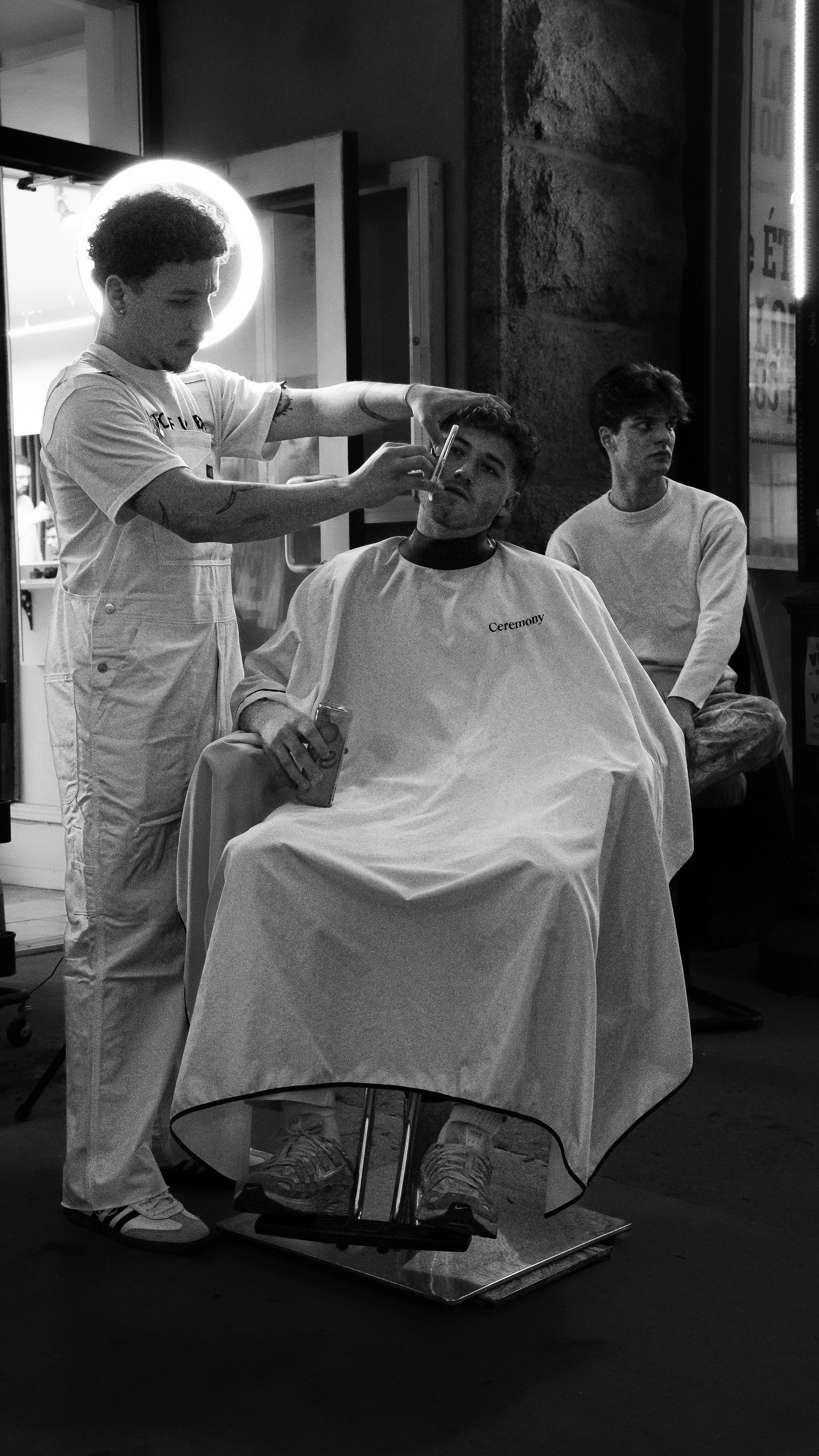 Un homme se fait couper les cheveux dans un salon de coiffure, une personne qui lui sert de client et une autre en arrière-plan.