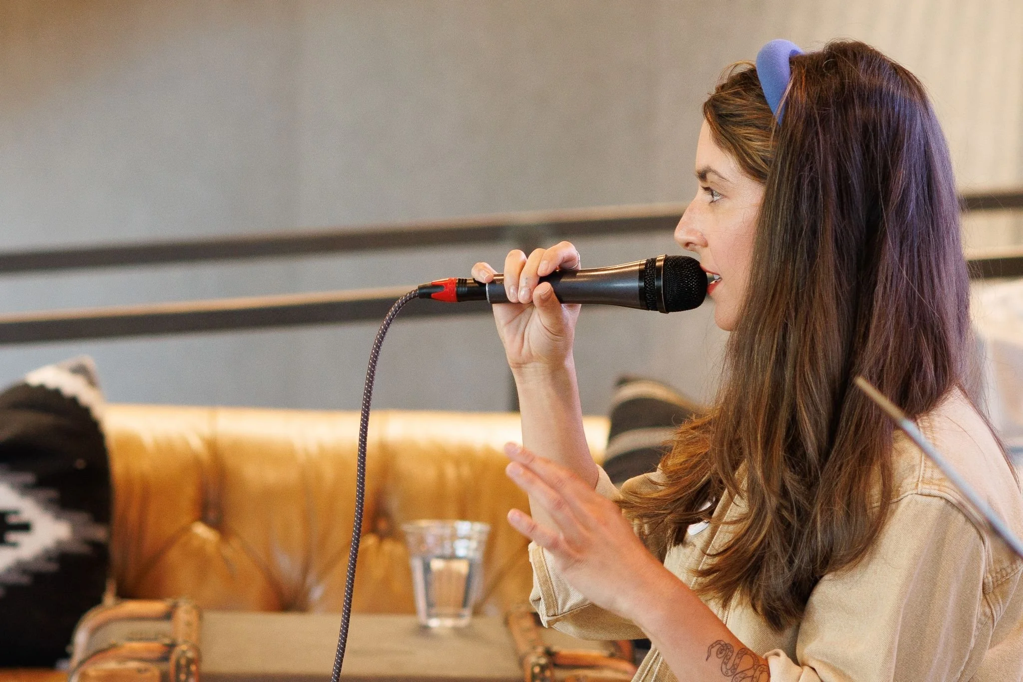 A woman with long brown hair and a blue headband singing into a black microphone in a cozy indoor setting.