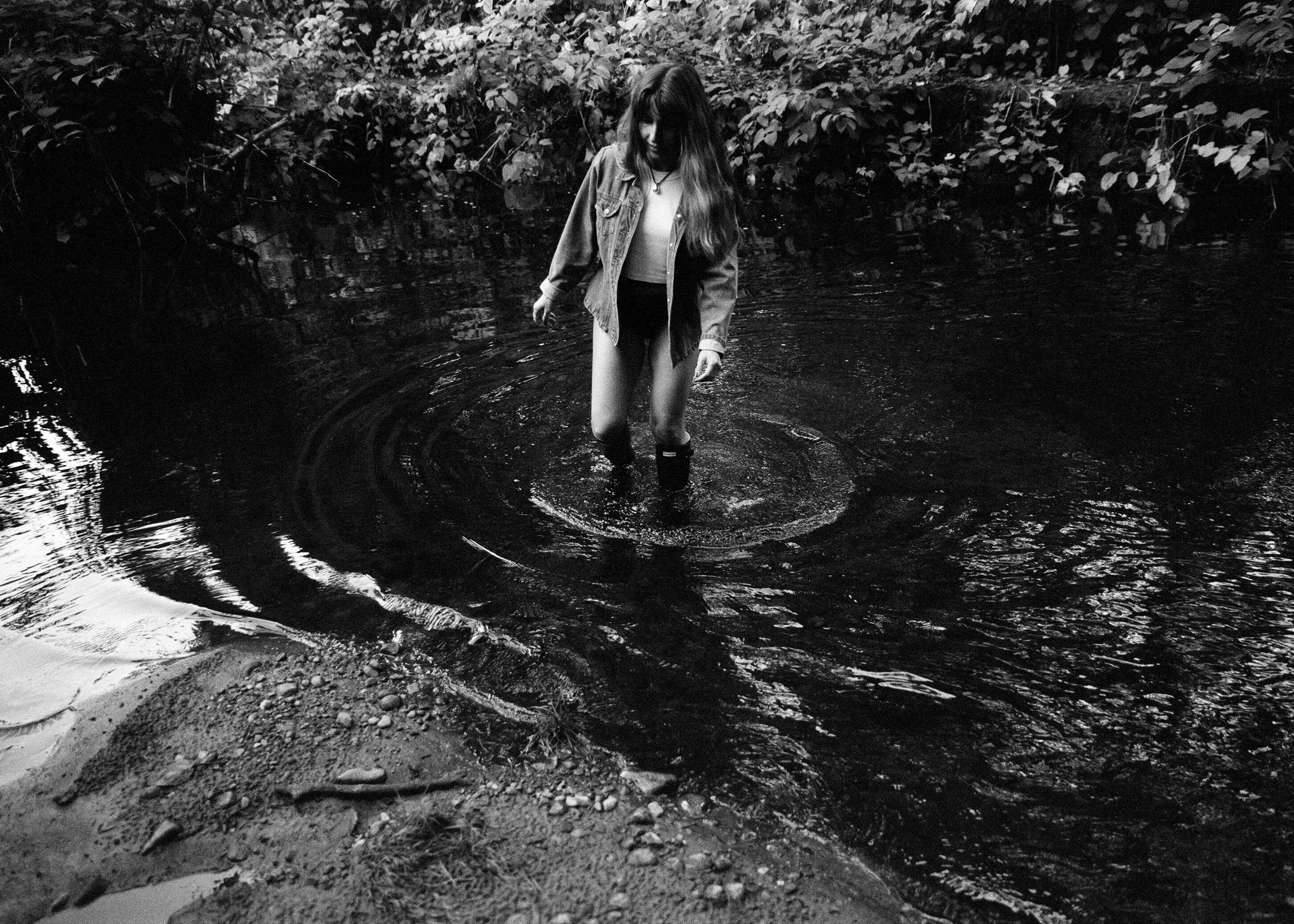 A girl with long hair wearing a jacket and boots standing in a shallow river surrounded by dense foliage