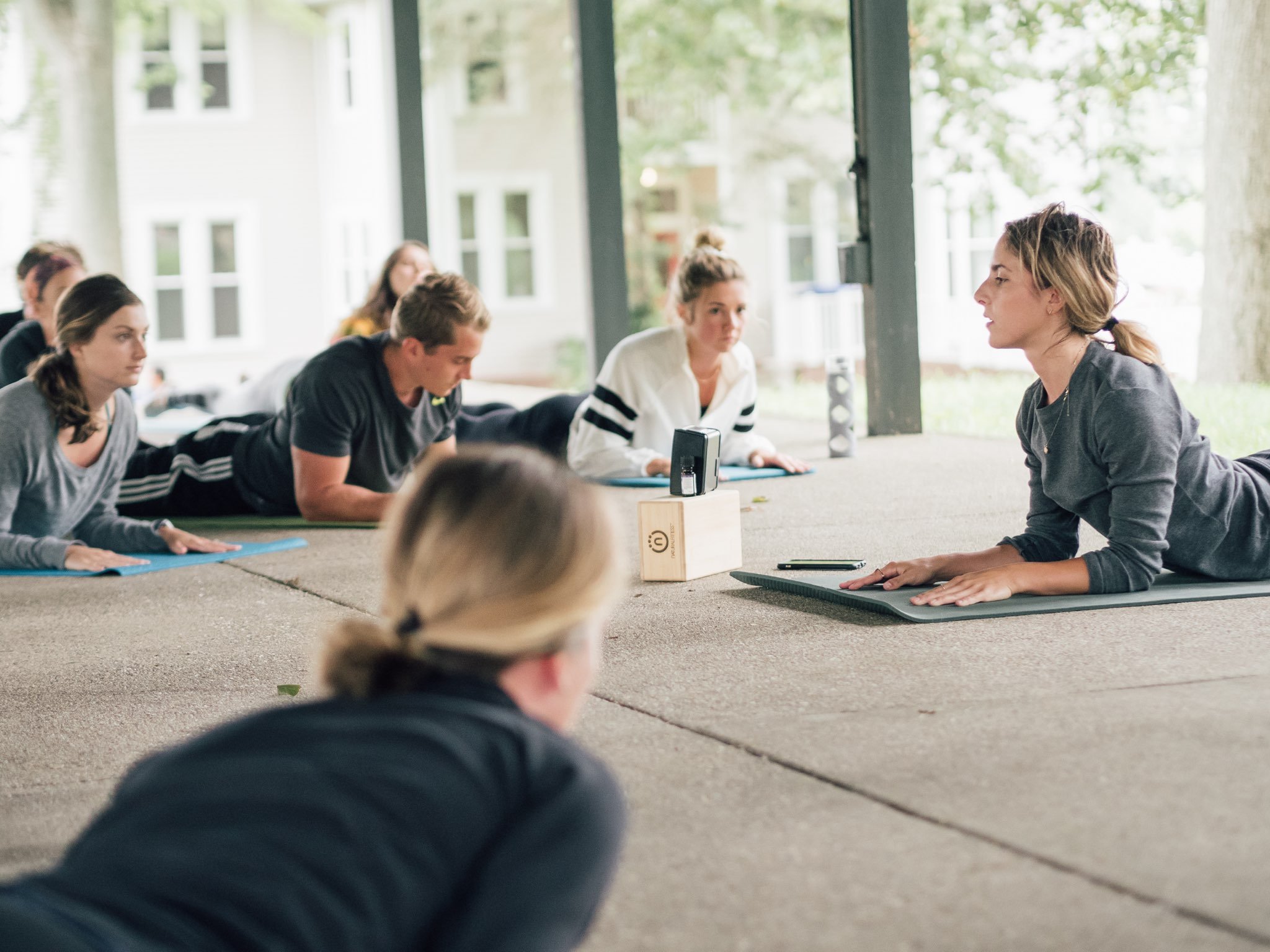 A group fitness instructor leading a yoga class outdoors under a pavilion, with participants lying on exercise mats in a prone position.