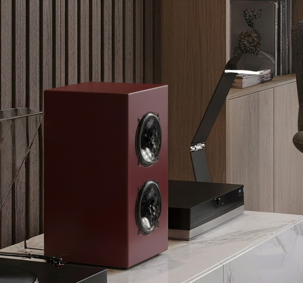 Altoparlante di colore rosso scuro su un mobile di marmo, con una lampada nera a LED e una mensola con libri e decorazioni di conchiglie in legno sullo sfondo.