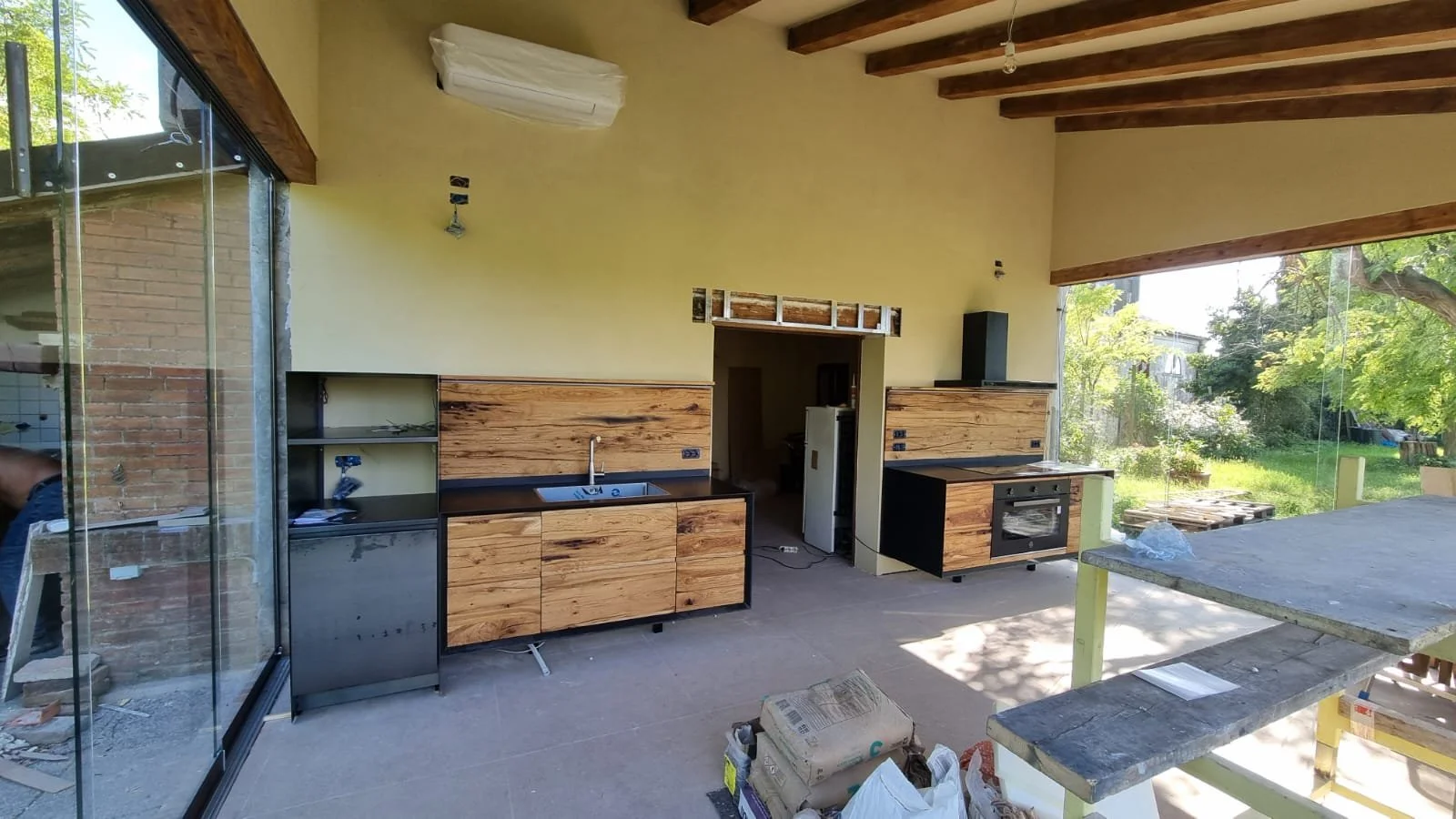 Interno di una cucina in costruzione con armadietti in legno e tubi, vista verso grandi finestre con giardino esterno