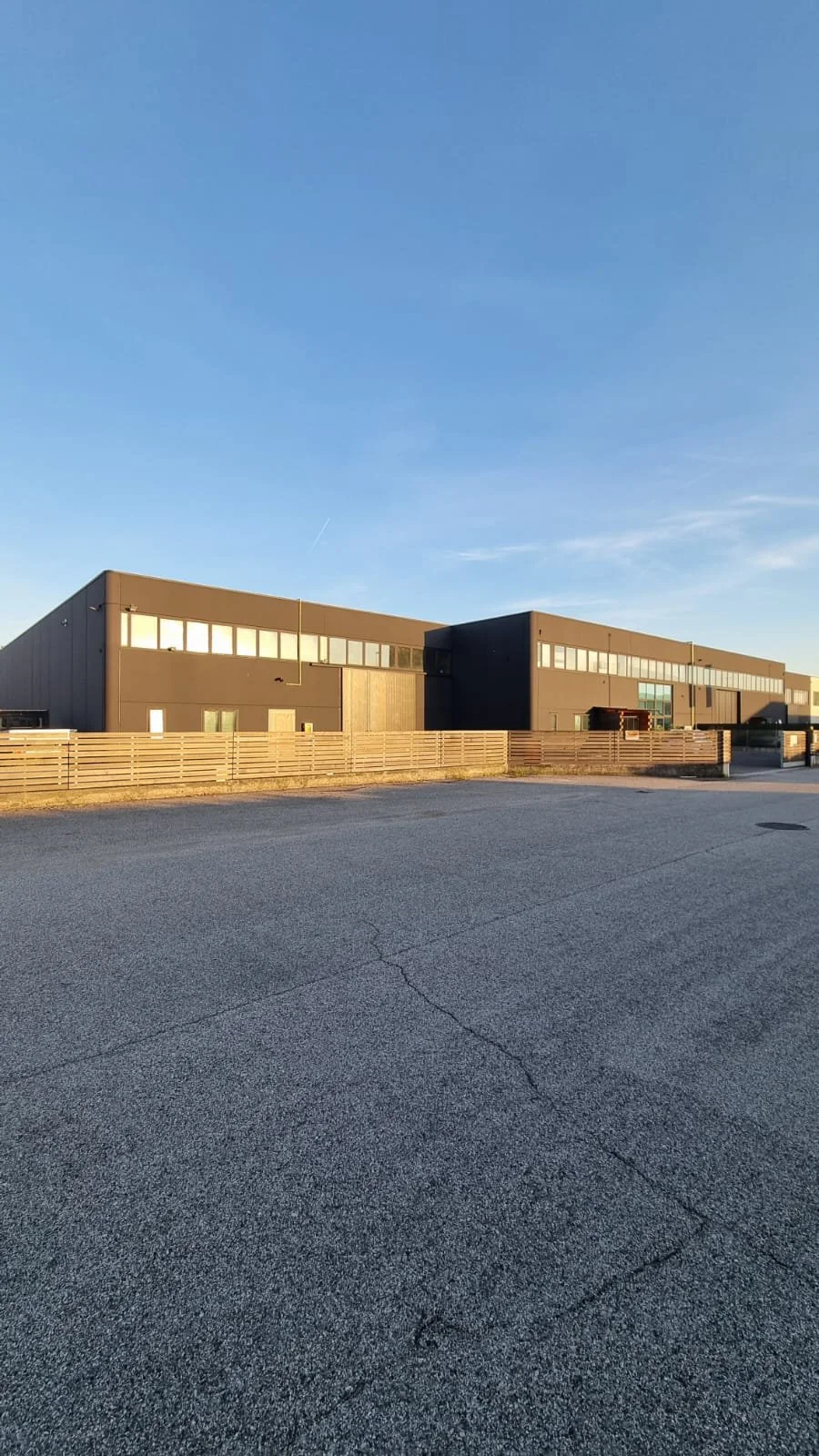 Edificio industrial moderno con pareti nere, finestre alte e un'area recintata in primo piano, sotto un cielo sereno con poche nuvole.
