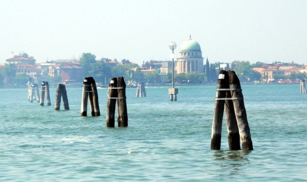 Elementi di una riva a Venezia con pali di legno nell'acqua e un edificio con cupola sullo sfondo.