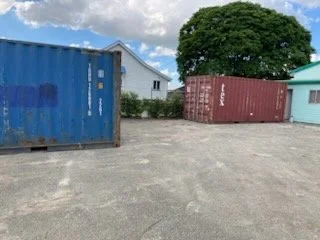 Two shipping containers, one blue and one red, placed on an open gravel lot with a few houses and a large tree in the background.