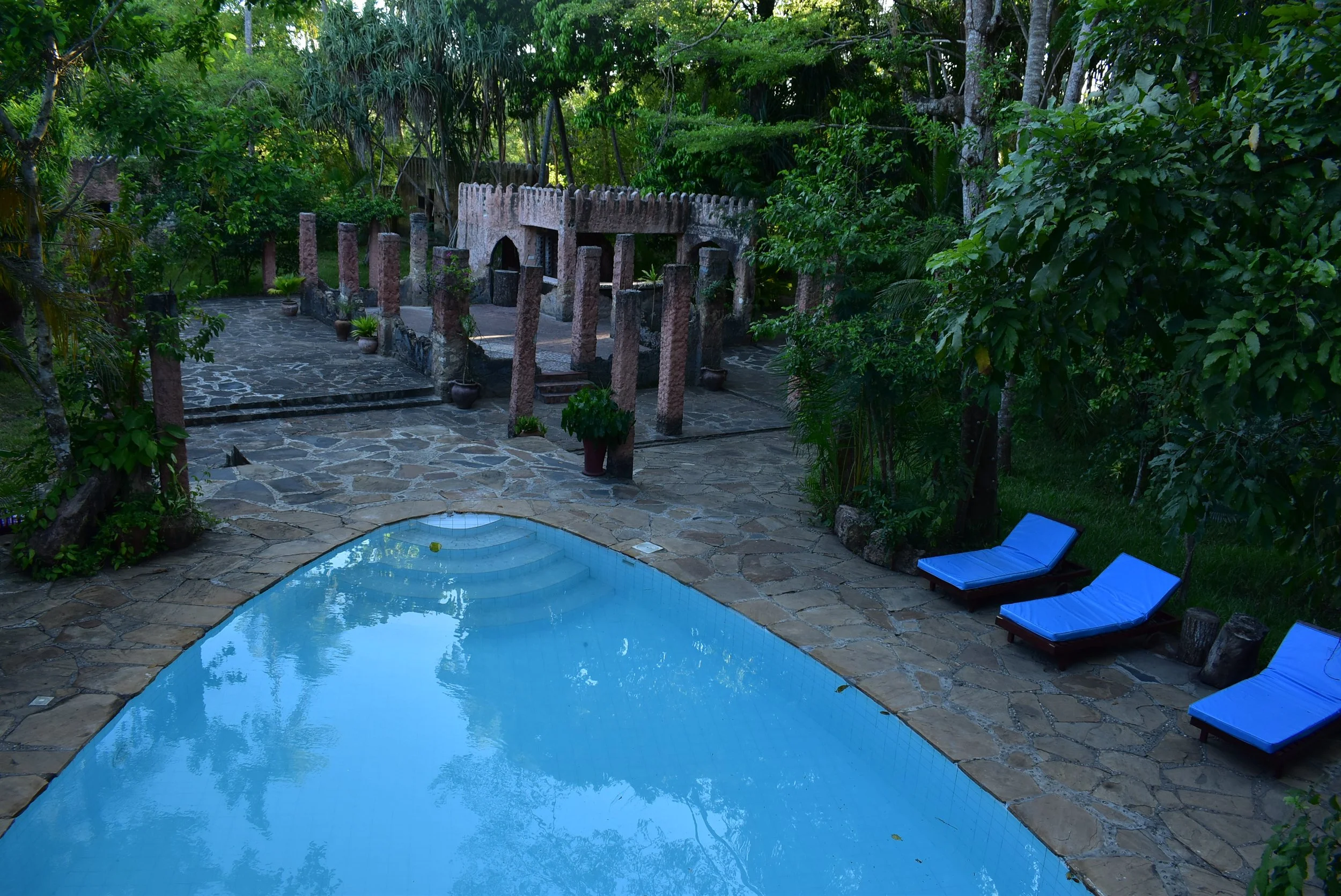 A tranquil outdoor pool area surrounded by lush green trees and plants, with three blue lounge chairs on a stone-paved deck and a decorative stone structure with columns and arches in the background. Where Utopia Experience: Culture Pool Fest