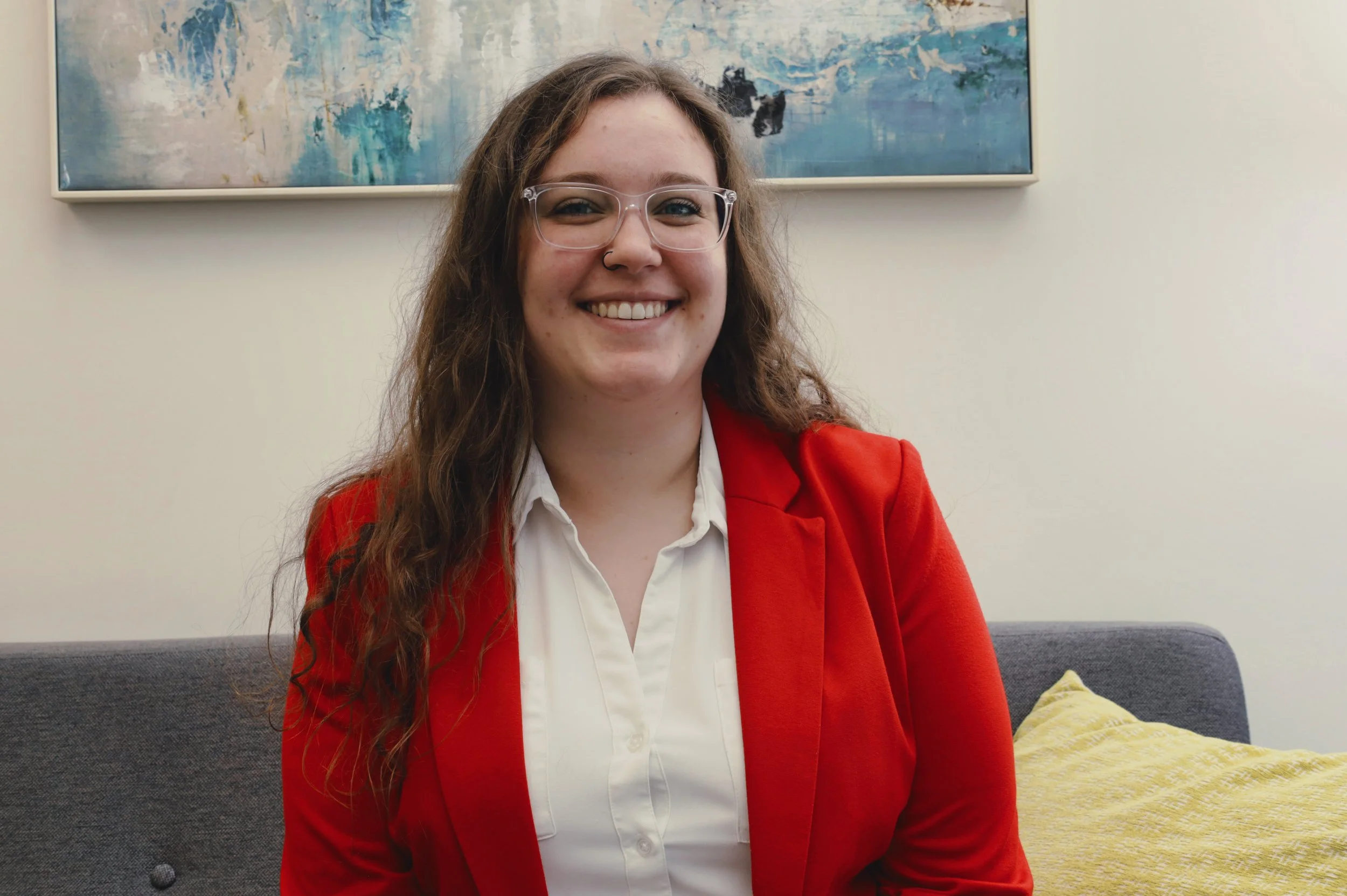 Close-up of a man with dark, curly hair, wearing clear round glasses and a nose ring. He has a beard and is dressed in a dark green sweater with a white shirt underneath. The background shows a blurred indoor setting with a plant.