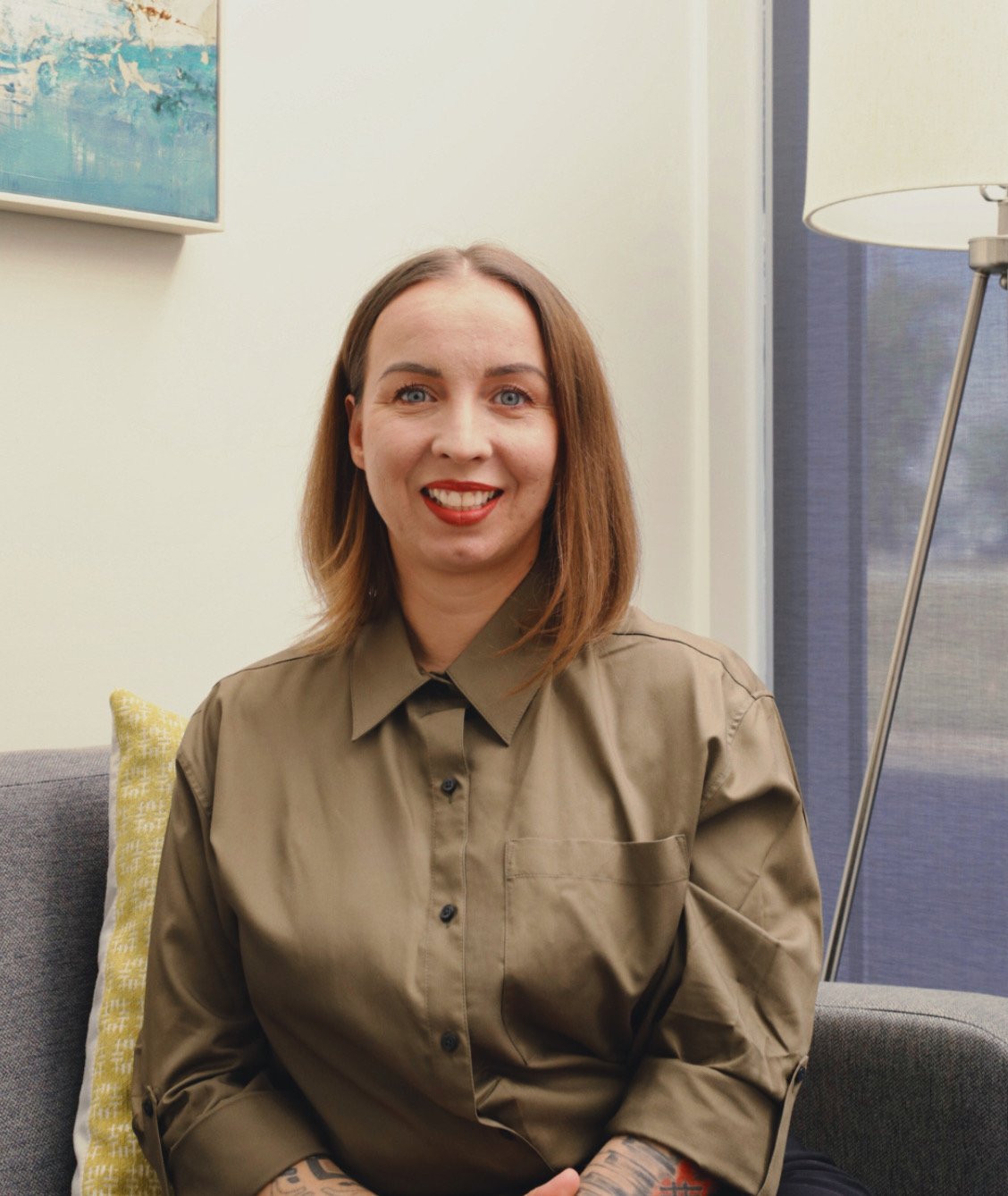 A woman with shoulder-length dark hair smiling outdoors with green plants in the background.