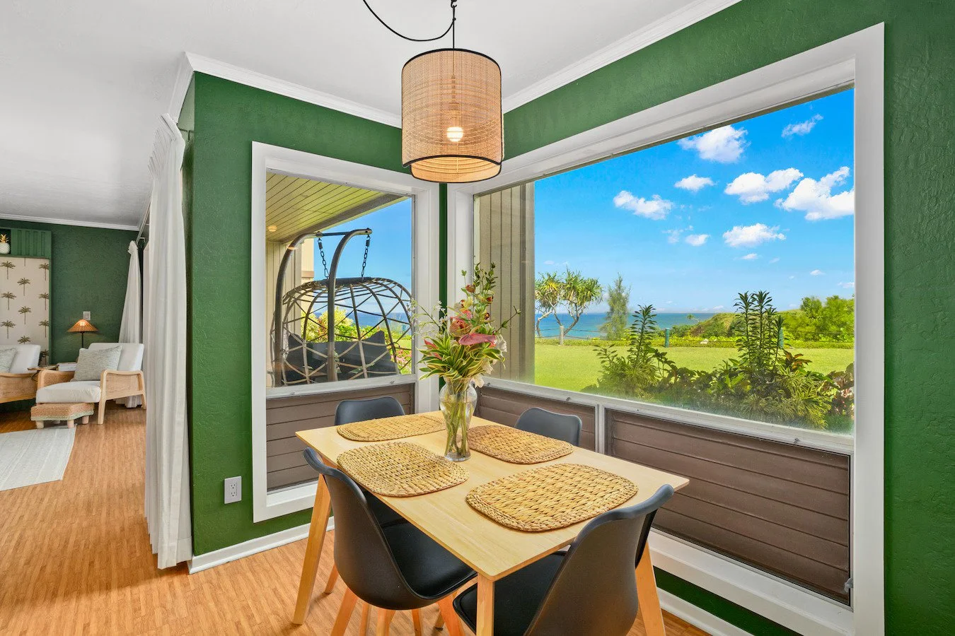 Dining area with a wooden table, four black chairs, and a vase with pink flowers. Large windows reveal a green yard, trees, and a blue sky with clouds, with a view of the ocean in the distance.