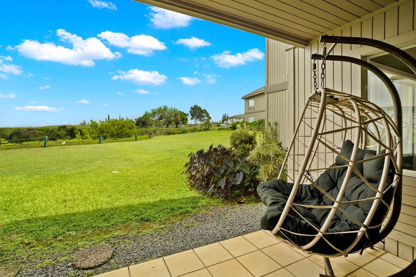 A patio porch with a hanging egg chair, black cushion, outdoors view of a green lawn, bushes, trees, and a blue sky with white clouds.