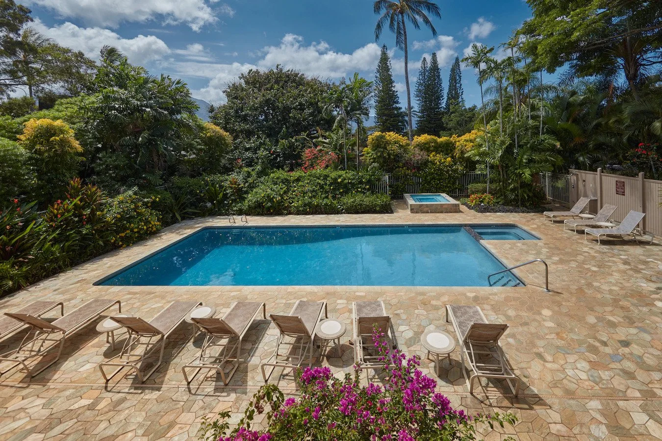 Outdoor swimming pool with surrounding lounge chairs, lush tropical trees and bushes, a hot tub, and sunny sky with some clouds.