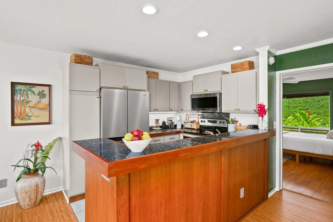 Kitchen with gray cabinets, a black granite countertop, a silver refrigerator, and a stove with a microwave above. There is a bowl of apples on the counter, a potted plant near the wall, and a pink flower in a vase on the bar. Part of a bedroom with 