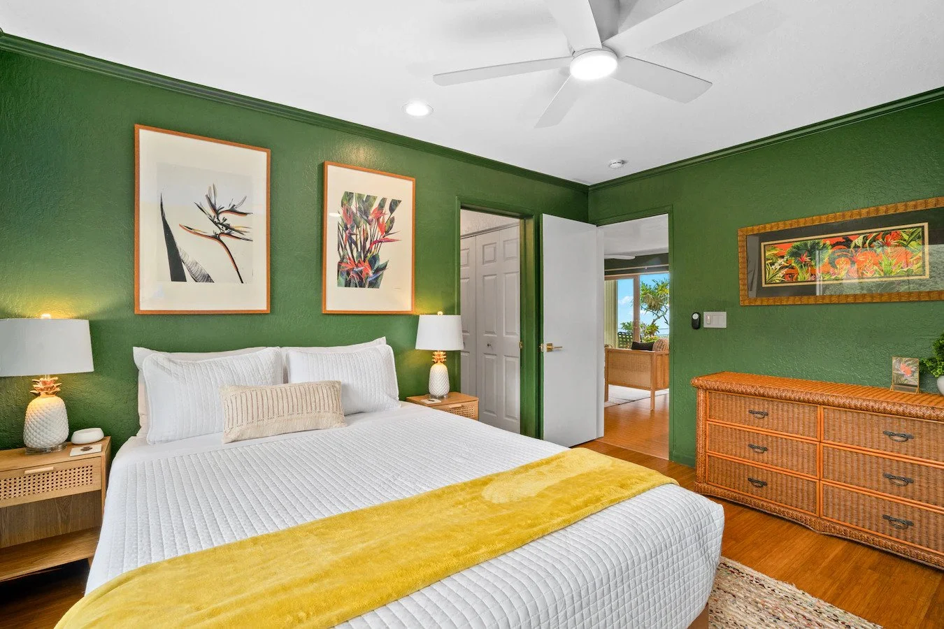 A bedroom with green walls, a white bed with pillows, and wooden furniture, including a dresser and nightstands with lamps. Artwork hangs above the bed and on the wall, with a door opening to the living room space showing trees and blue sky.
