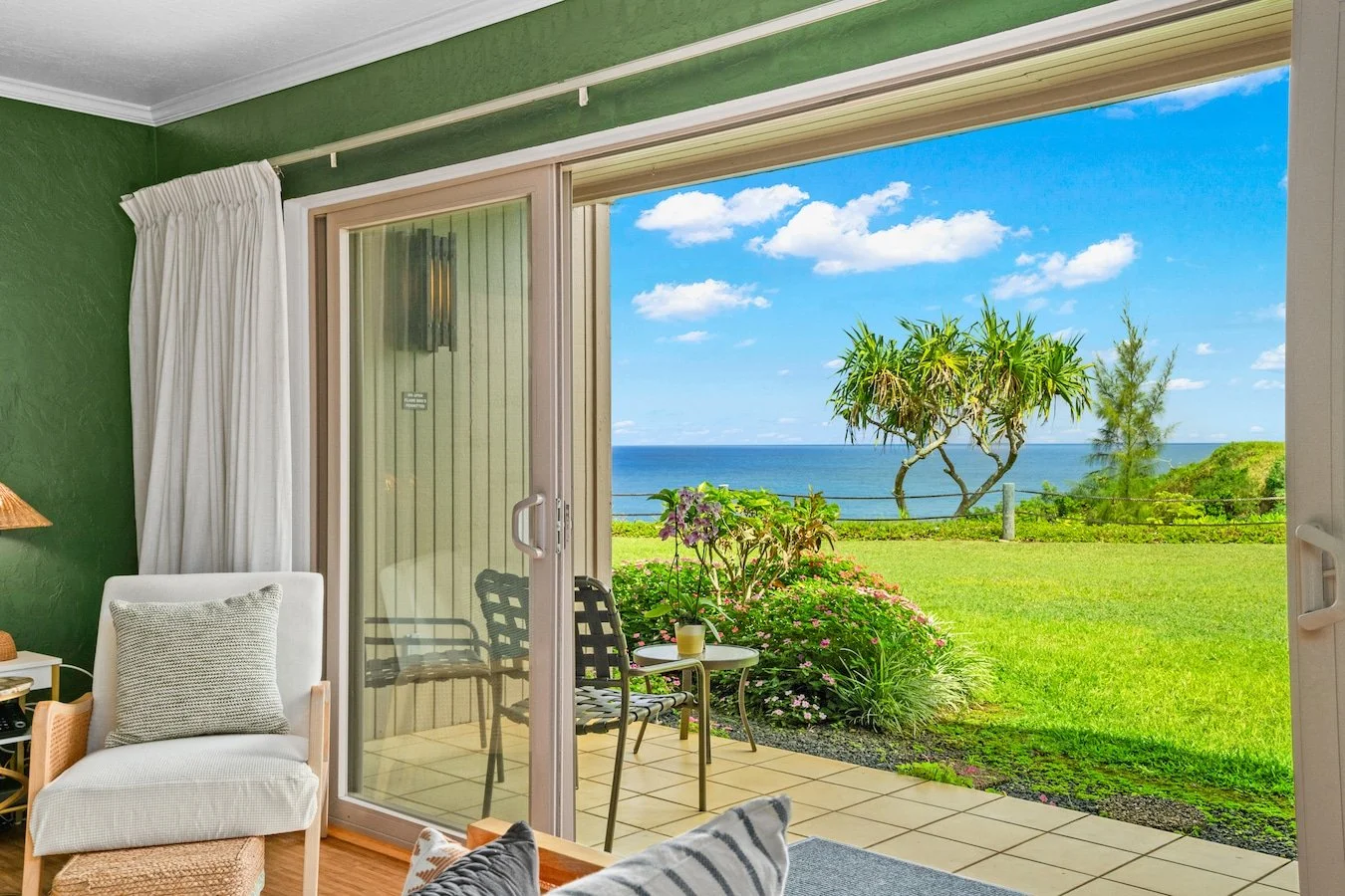 Living room with a sliding glass door opening to a patio and view of green grass, trees, and ocean under blue sky with clouds.