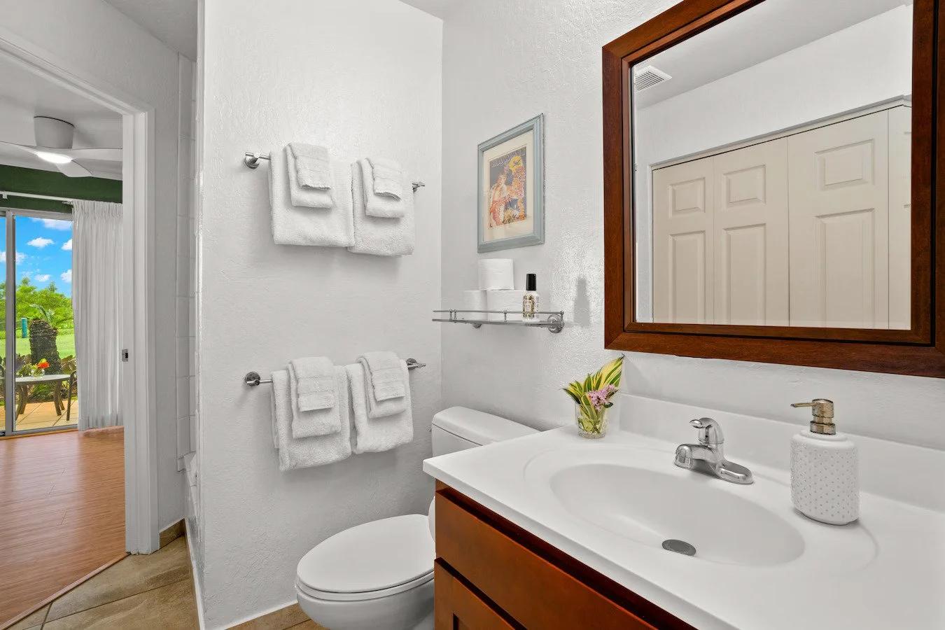 Bathroom with white walls, wooden mirror frame, white sink, and towels, with a view of a patio and green outdoor scenery through the doorway.