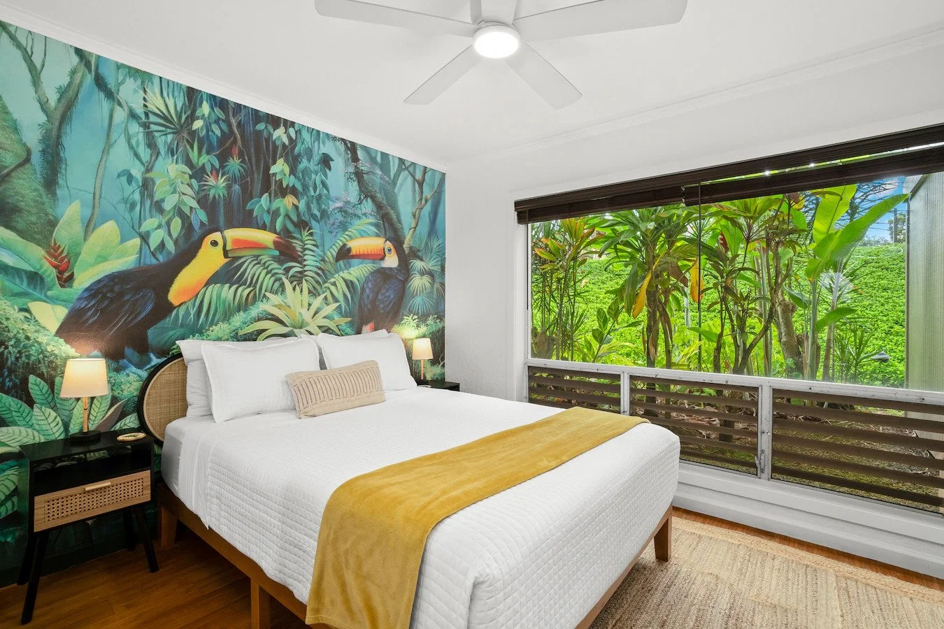 A bedroom featuring a mural of tropical birds with lush jungle foliage, a large window with lush greenery outside, a white bed with decorative pillows, bedside lamps, and a wooden floor.