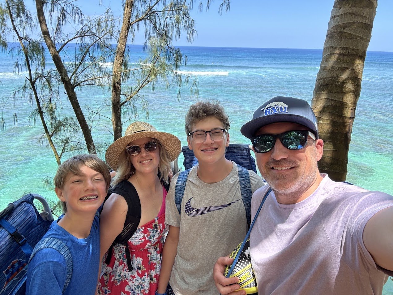 Family of four taking a selfie on a tropical beach with clear turquoise water, palm trees, and a blue ocean background, wearing casual summer clothing and accessories.