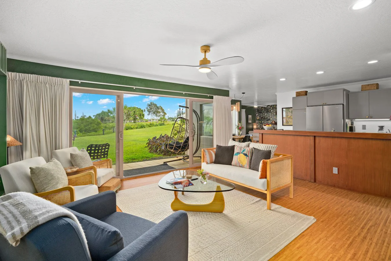 Living room with large sliding glass door leading to green outdoor space, white and wicker furniture, glass coffee table, hanging chair, and kitchen area in the background.