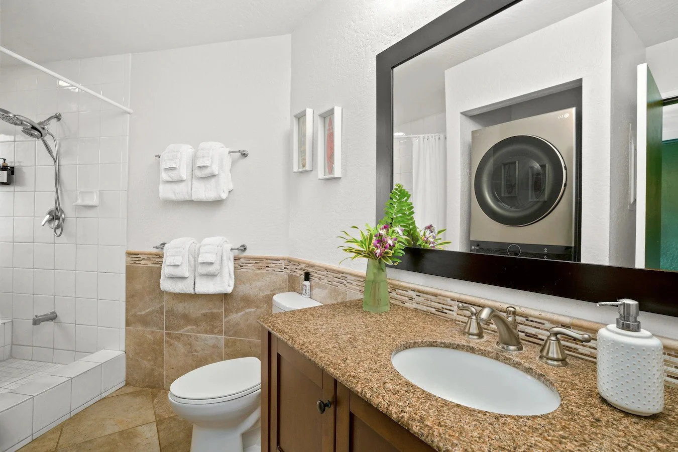 A bathroom with a shower area, toilet, vanity with granite countertop, a large mirror, and laundry appliances reflected in the mirror.