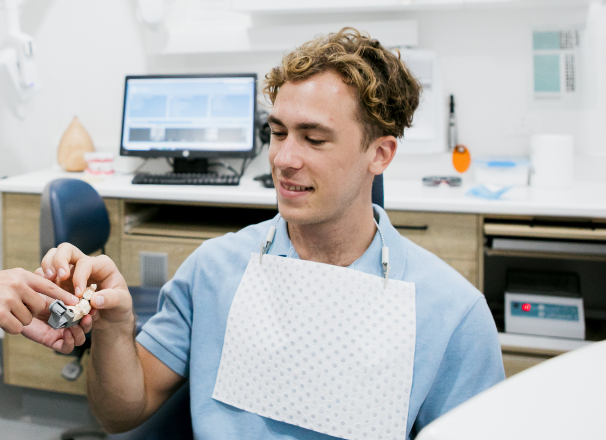 A young man with curly hair in a dental clinic, holding a dental model, smiling with a bib over his shirt.
