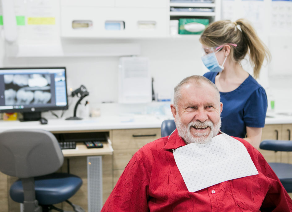 An older man sitting in a dental chair with a bib, smiling, while a female dental hygienist or dentist wearing a mask, glasses, and scrubs examines him at Battery Hill dental office.
