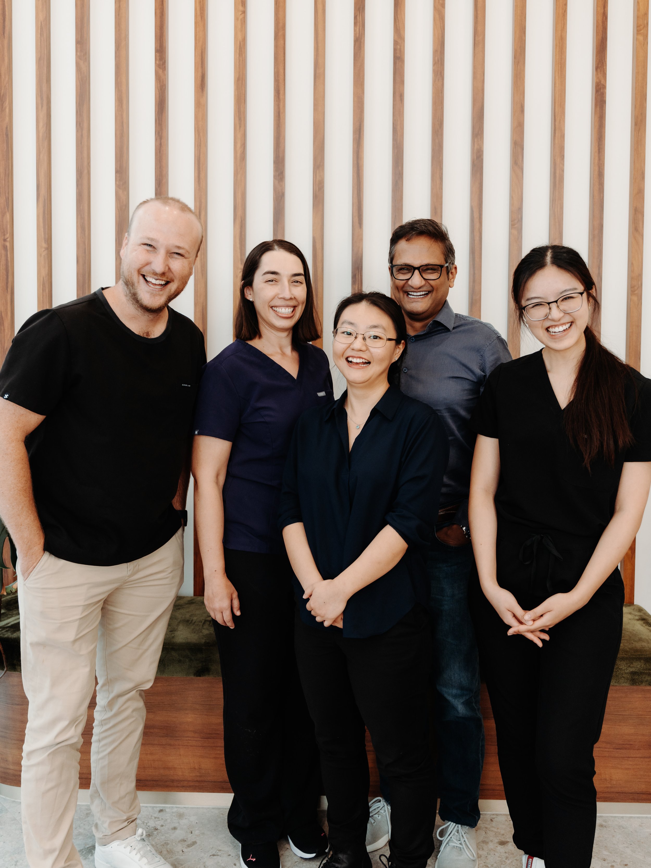 Group of five diverse people smiling and posing together indoors with wooden wall decor in the background.