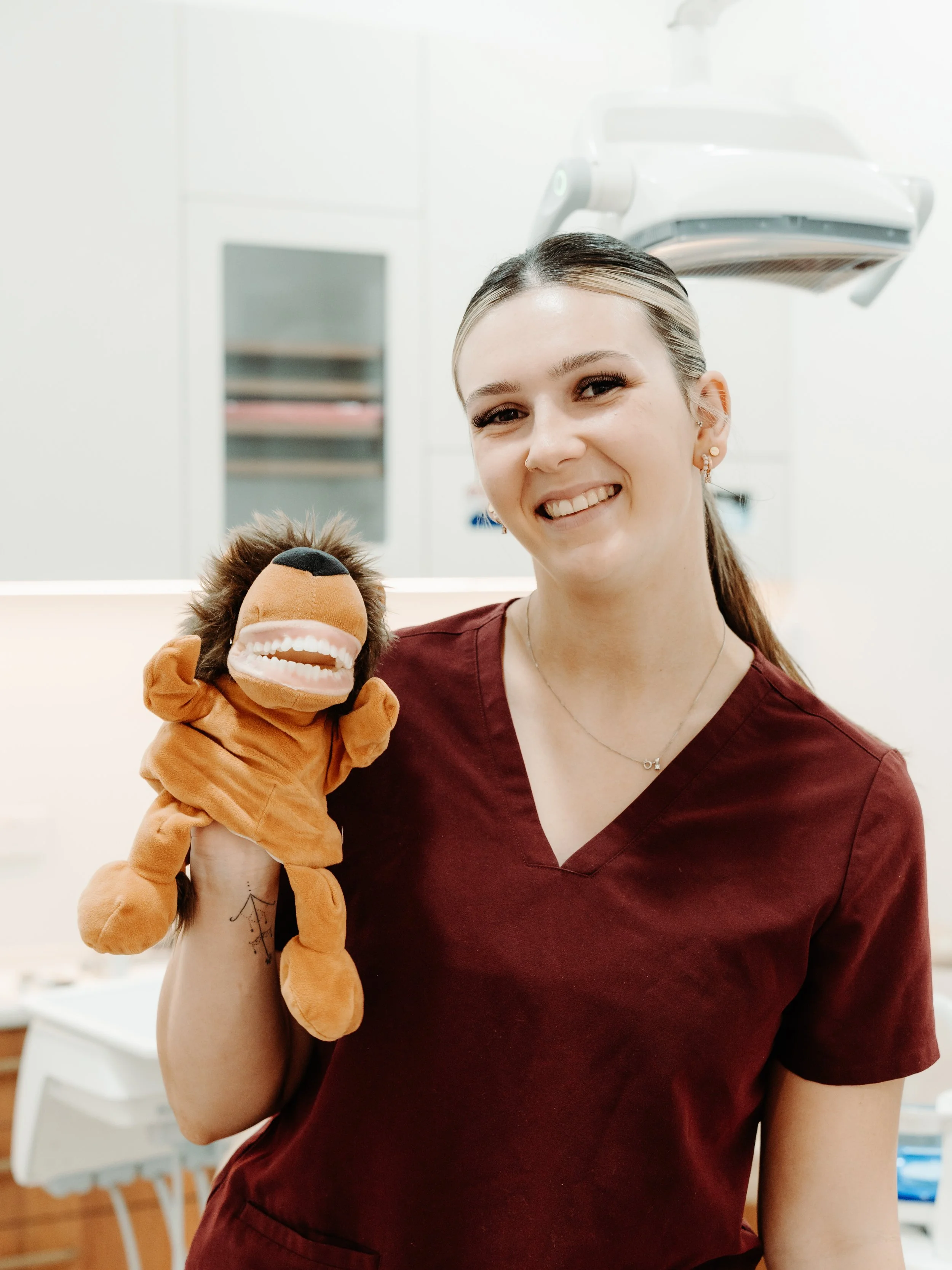 A smiling woman in a maroon medical uniform holding a lion plush toy at Wurtulla dental clinic.