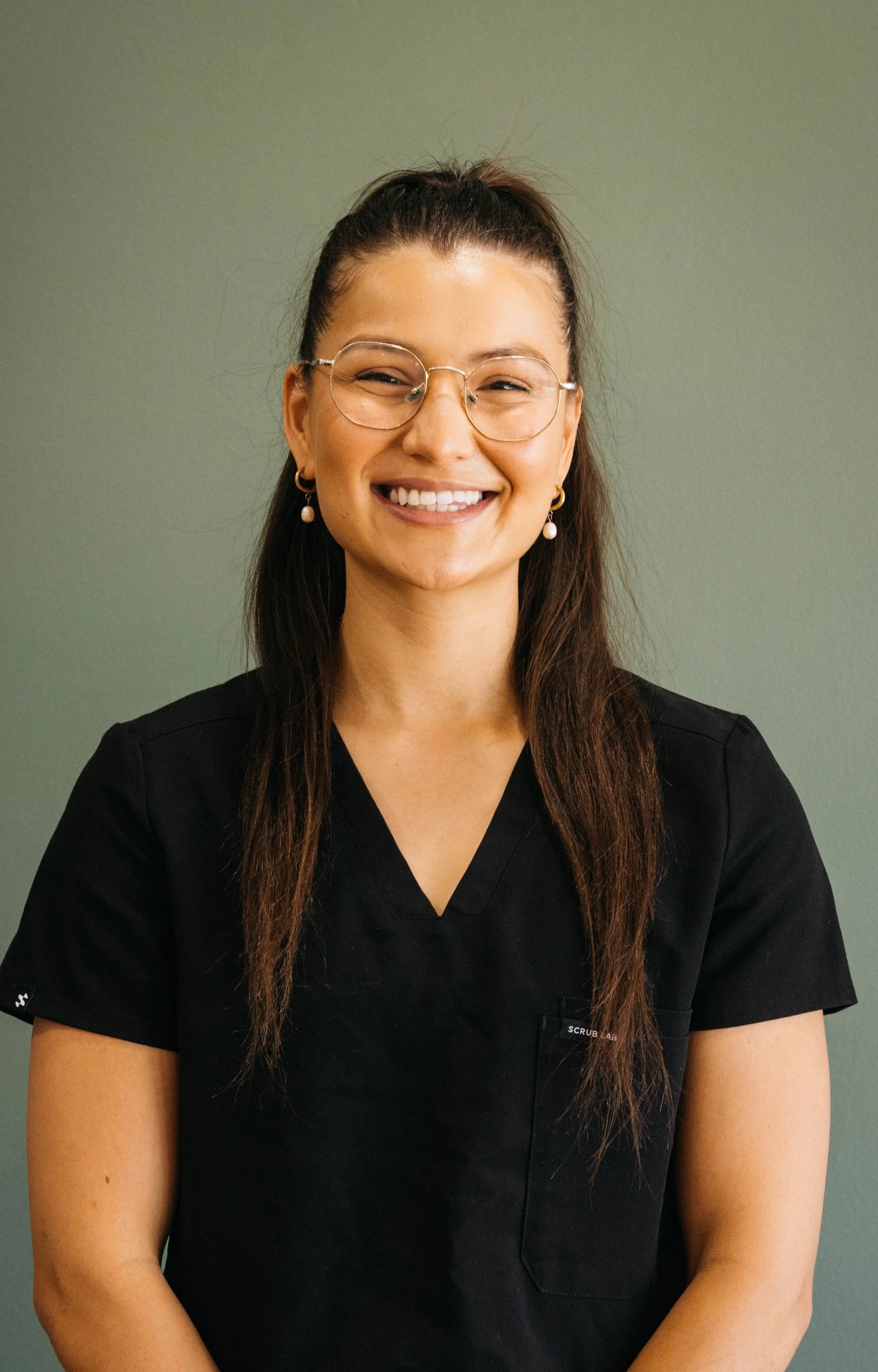 A woman in black scrubs smiling with glasses, earrings, and long dark hair against a muted green background.