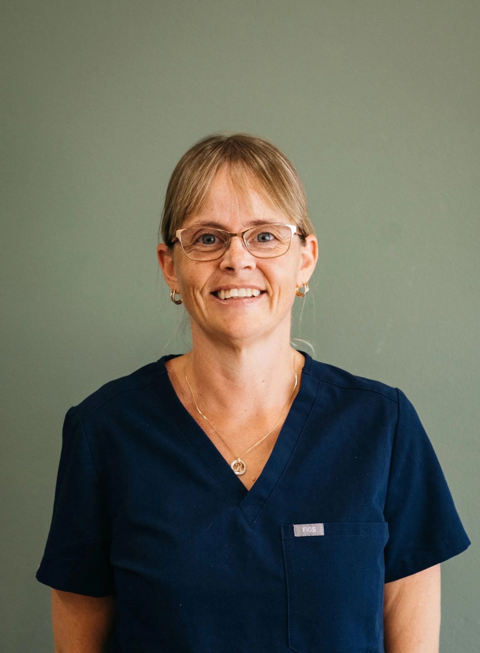 A woman in navy scrubs with glasses, earrings, and a necklace, smiling against a green background.