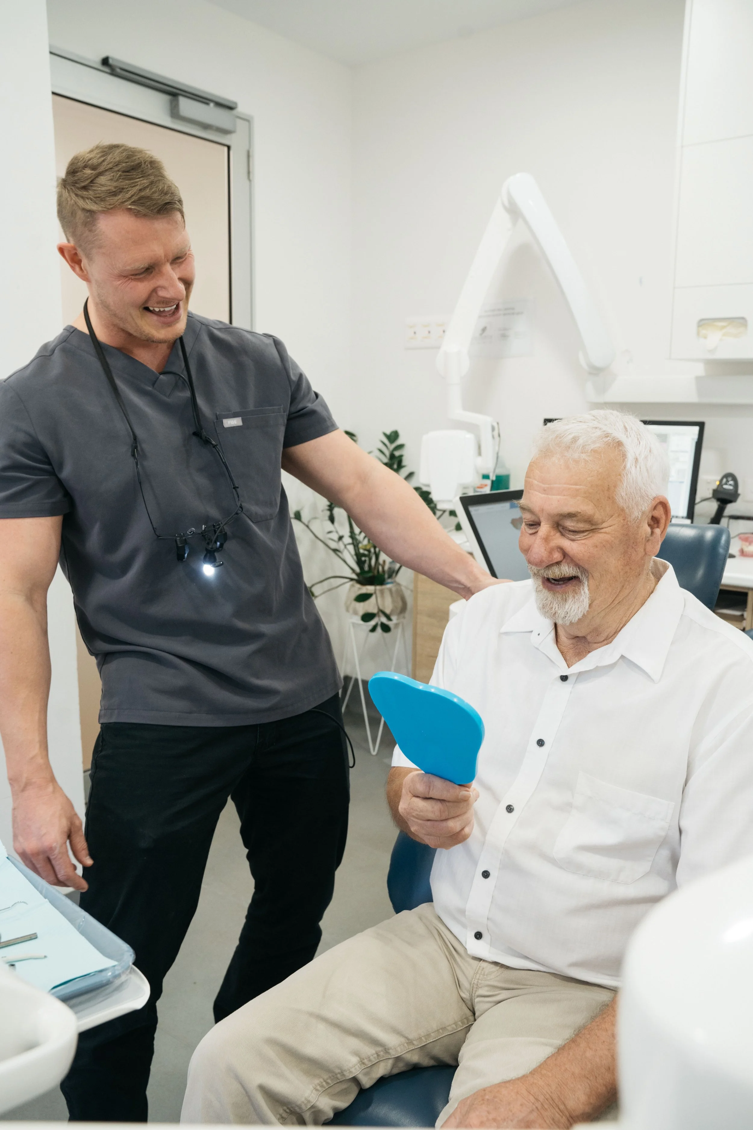 A dentist or dental hygienist with a magnifying headset smiling and talking to a senior male patient in a dental clinic. The patient is holding a small mirror and smiling.