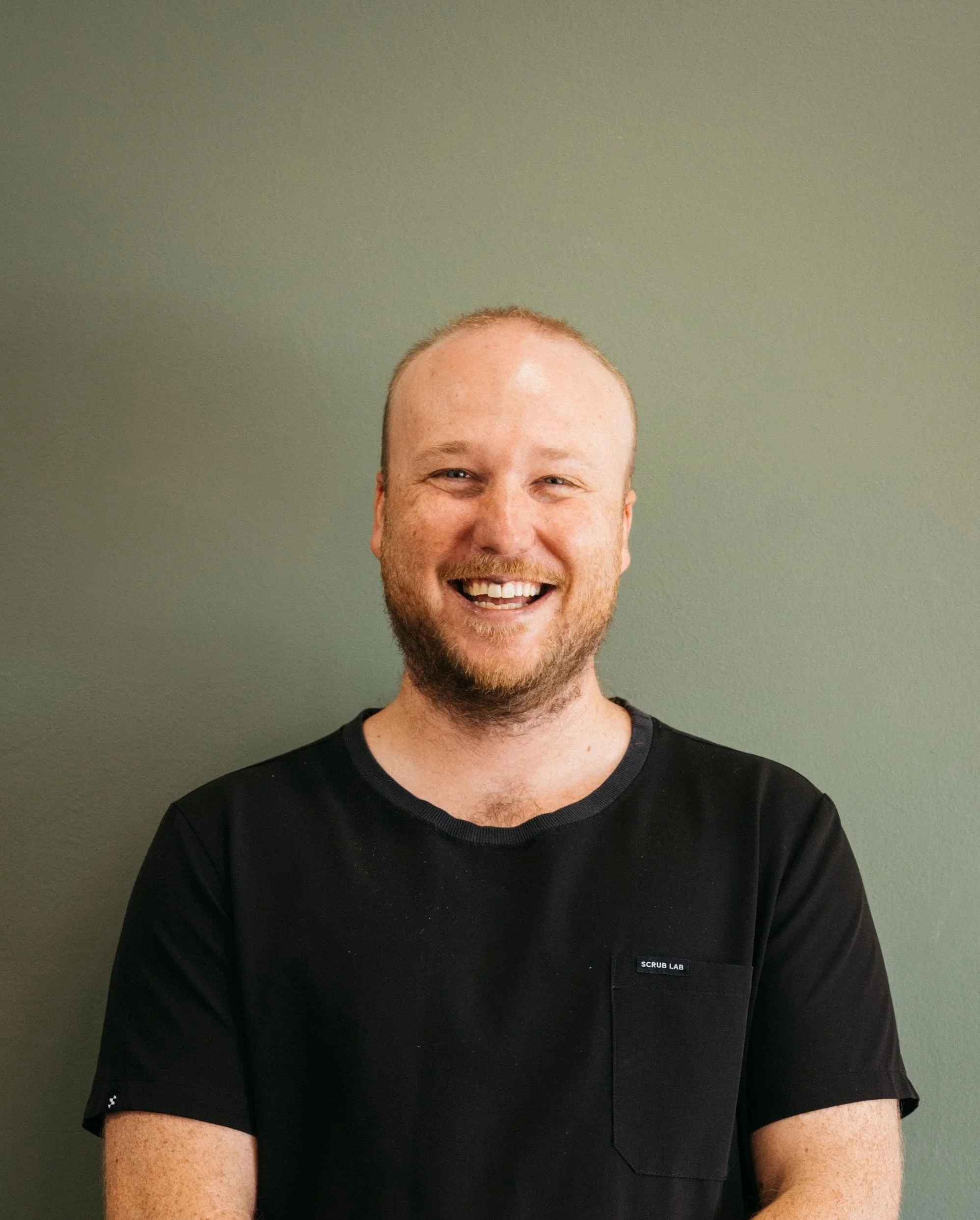 Smiling man with a beard and short hair, wearing a black shirt, standing against a green wall.