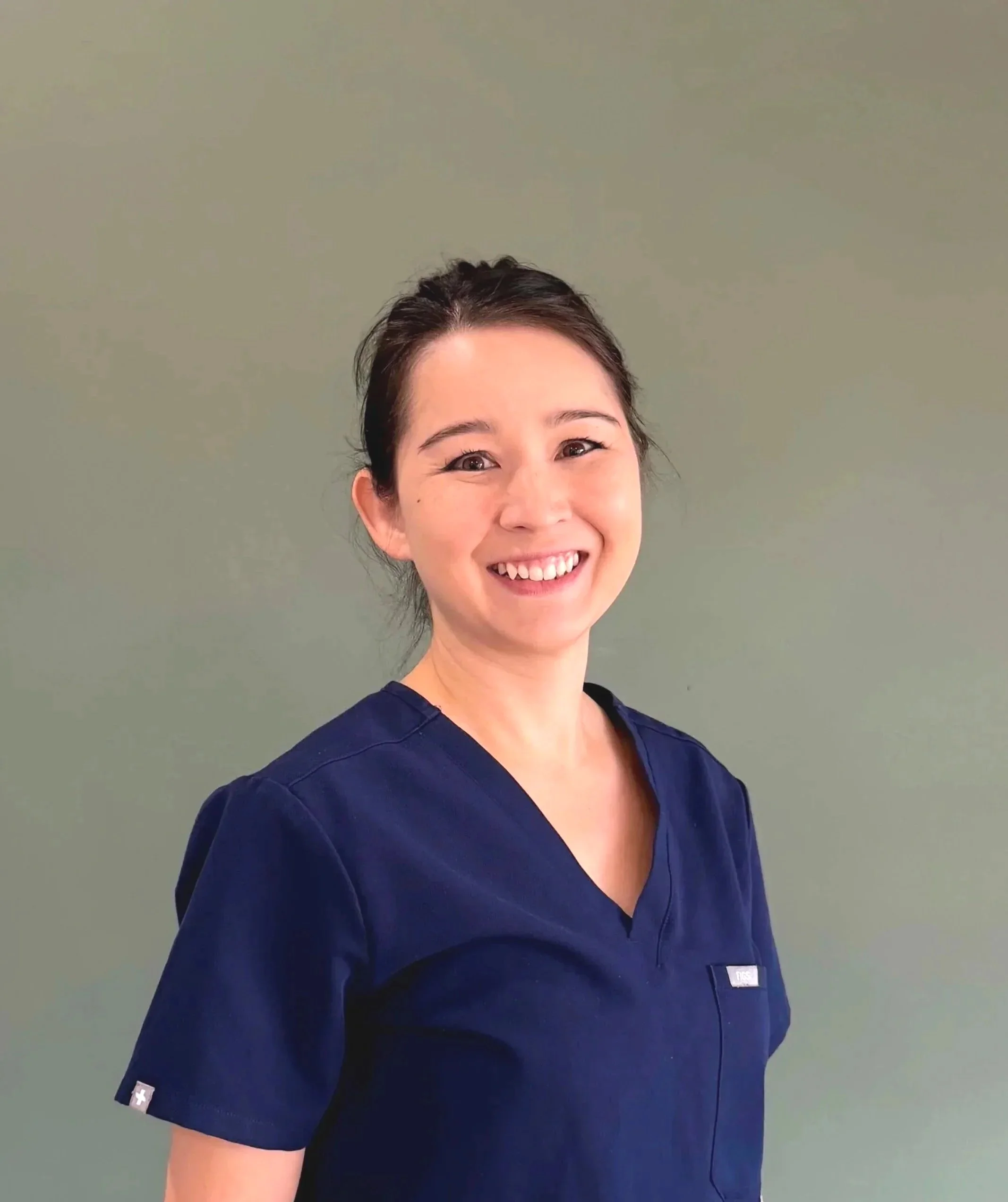 A woman smiling, wearing navy blue scrubs, standing against a plain, muted green background.