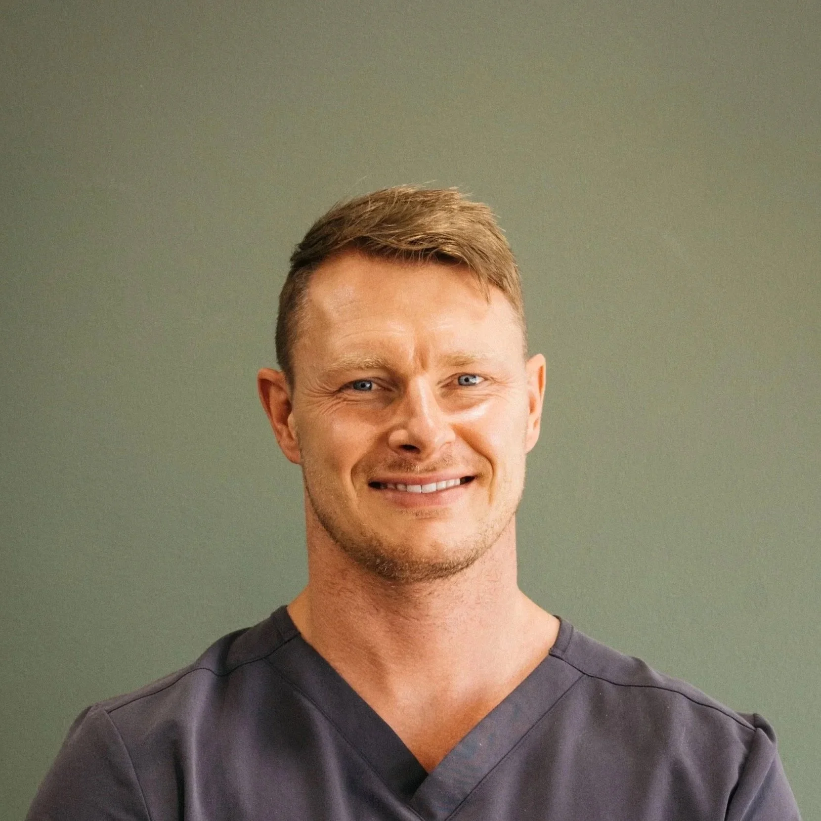 Portrait of a smiling man with short brown hair wearing dark scrubs against a green background.