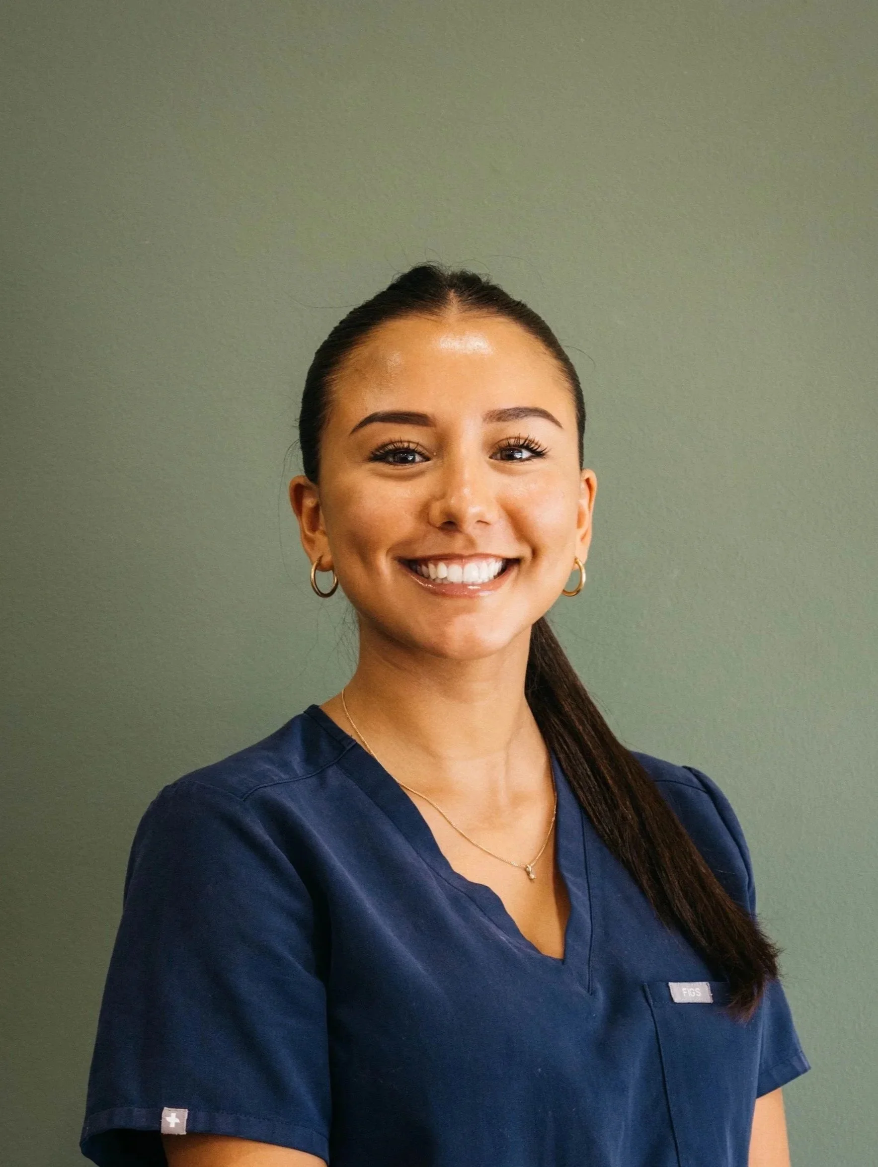 A smiling woman wearing navy scrubs, standing against a plain gray wall.