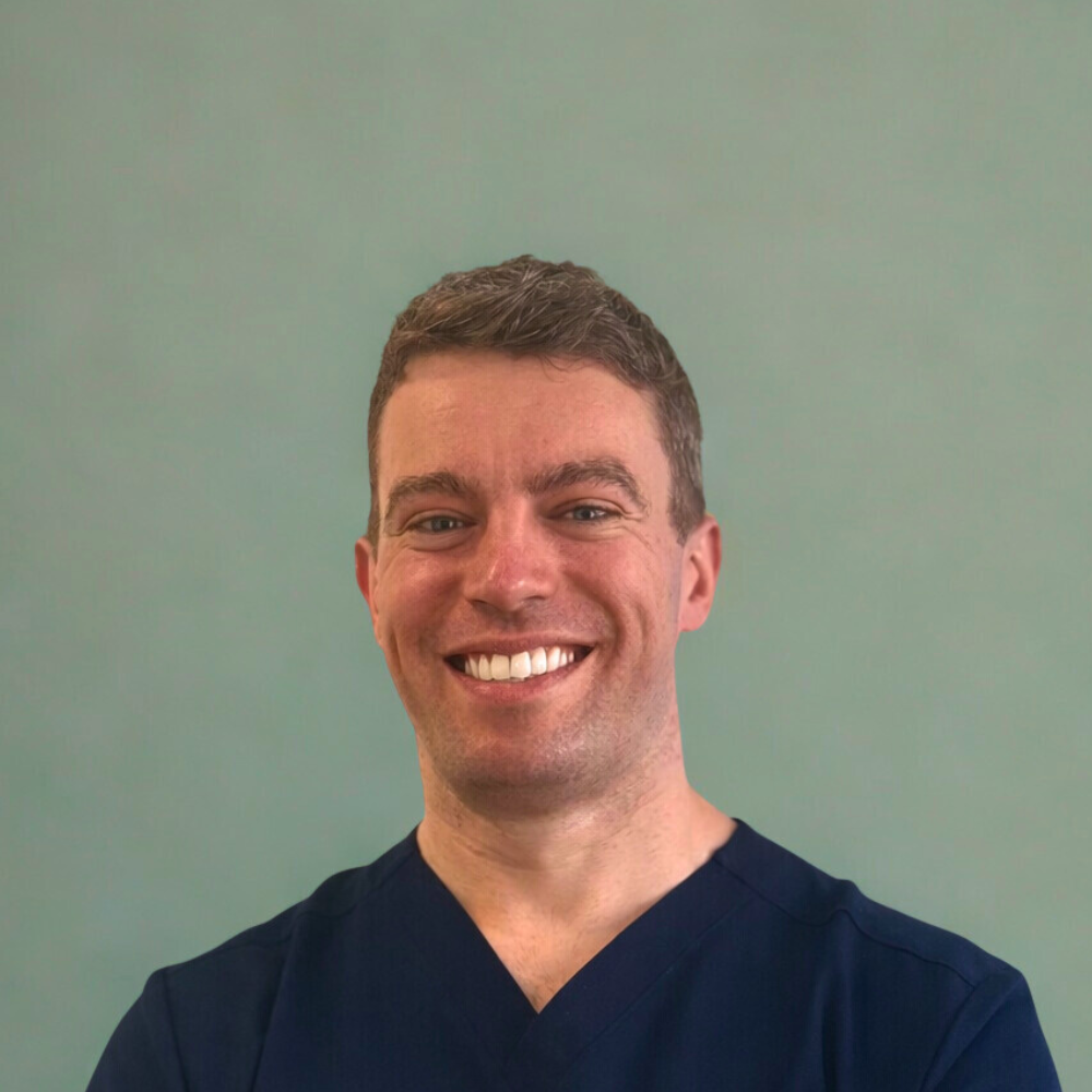 A smiling man with short brown hair in a navy scrub top standing against a plain green wall.