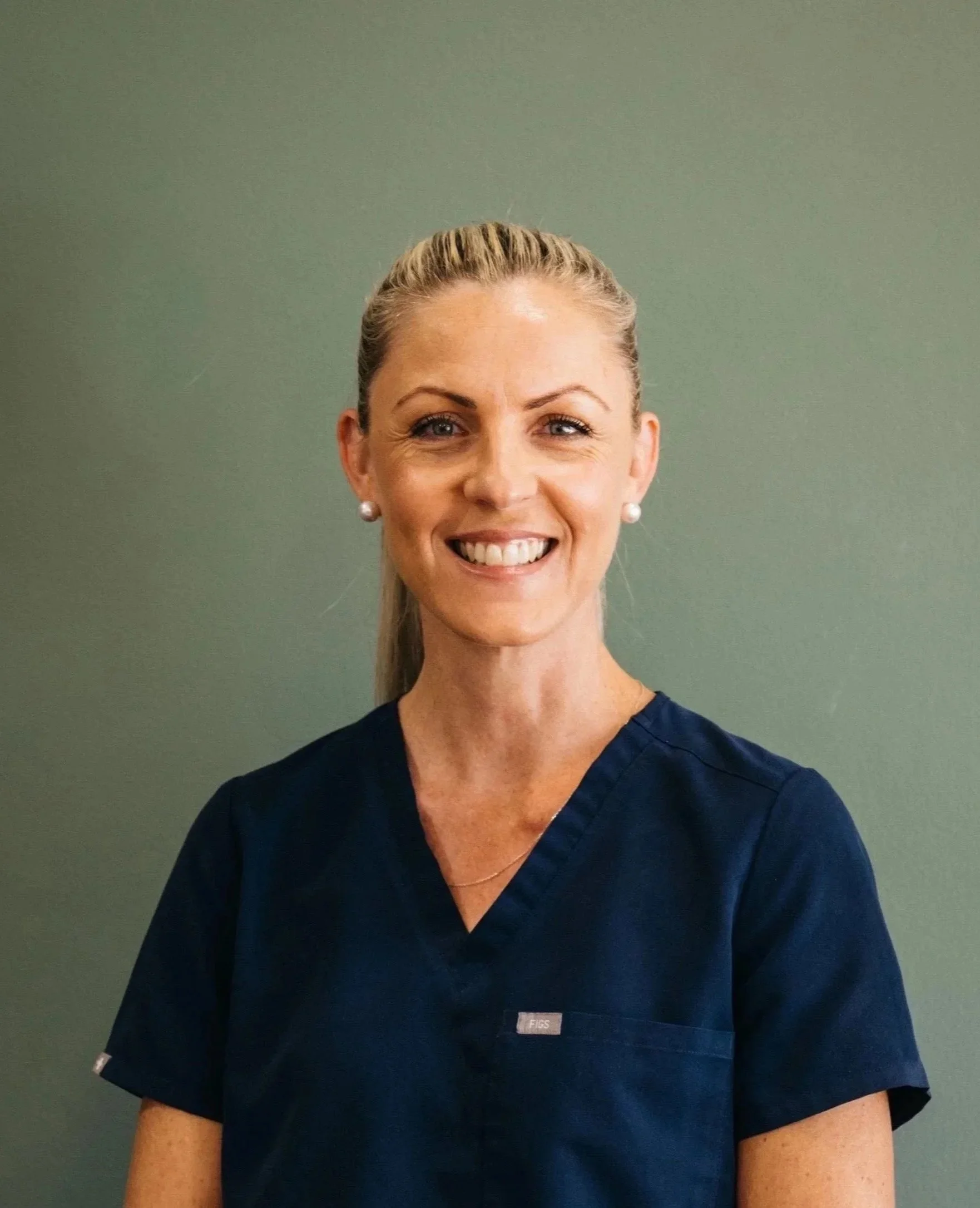 A smiling woman with blonde hair tied back, wearing pearl earrings and navy blue scrubs, standing against a green wall.