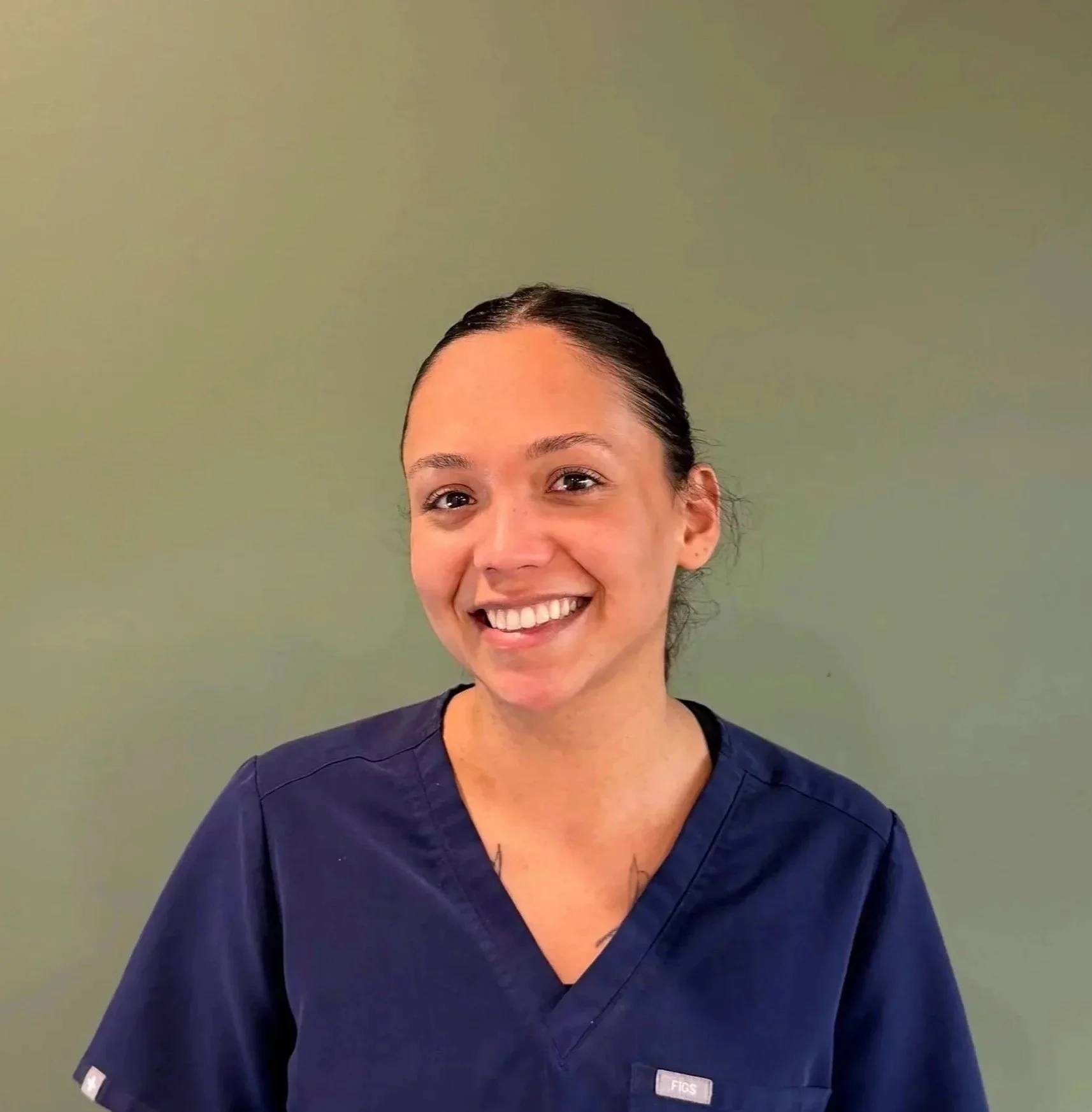A smiling woman in navy scrubs standing against a plain light green wall.