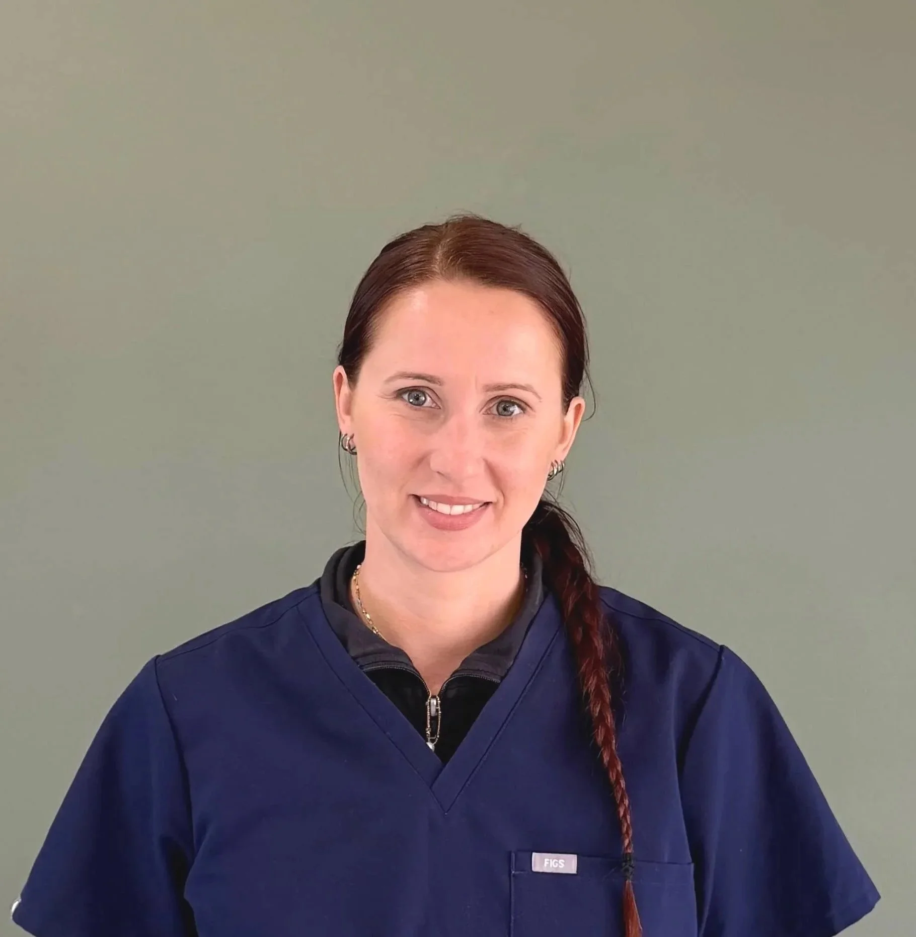 A woman with long red hair in a braid, wearing navy scrubs and a black zip-up top, standing against a neutral background.