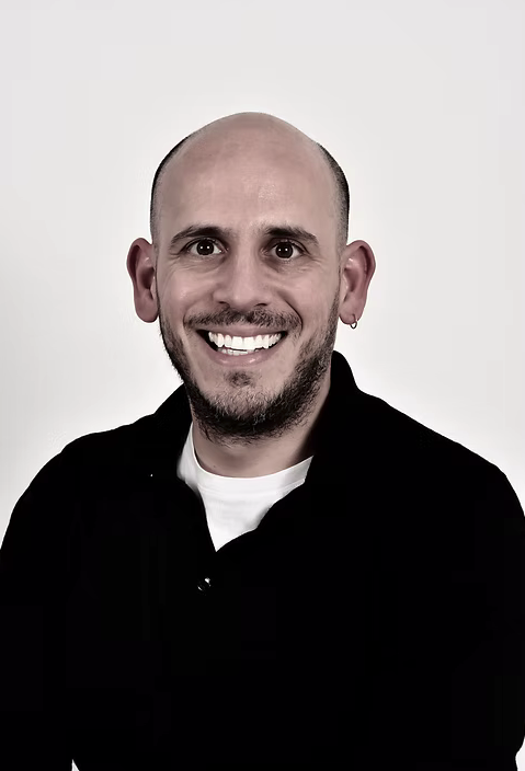 Smiling man with a bald head, beard, and earring in his left ear, wearing a black jacket and white shirt, against a plain white background.