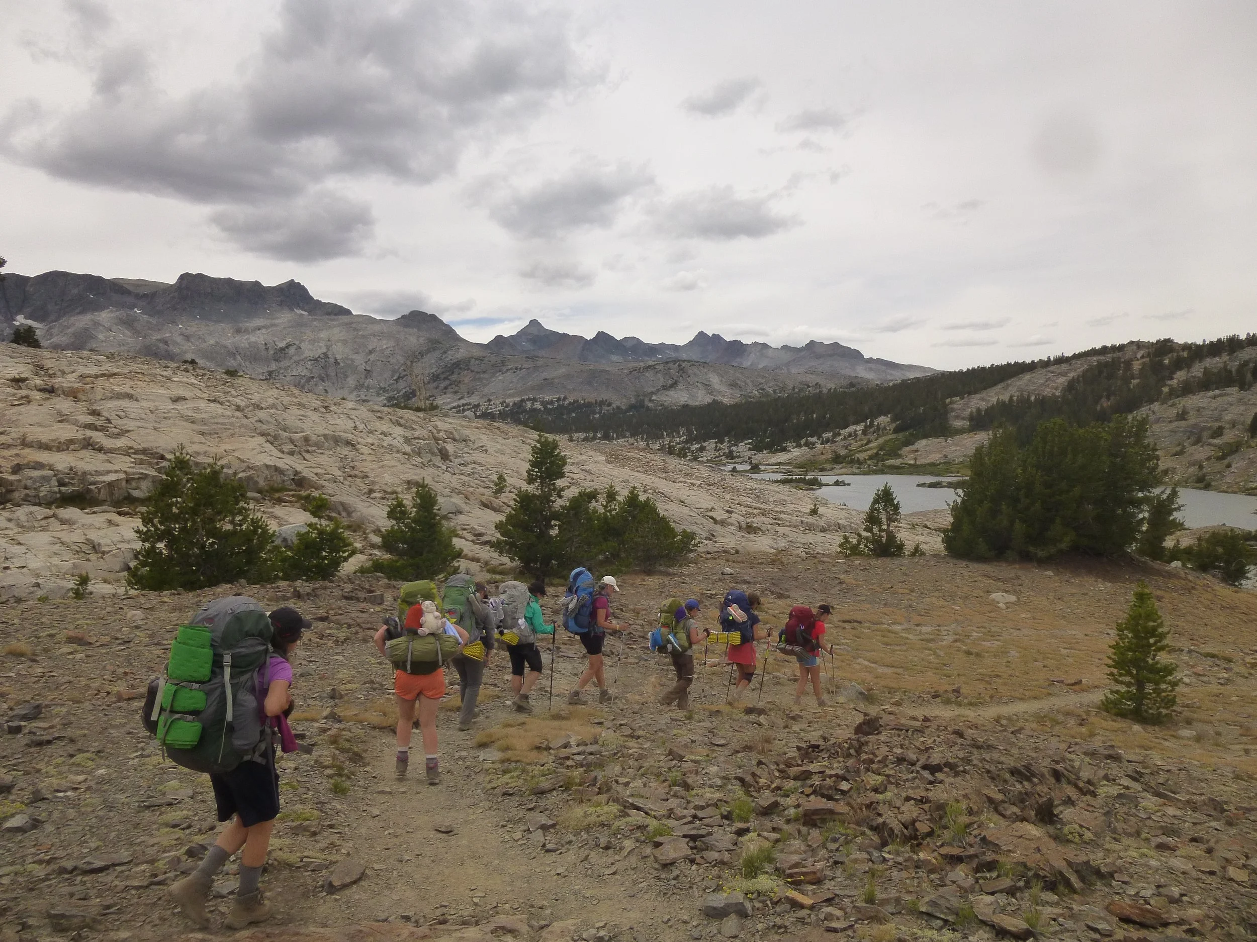 A group of Girl Scout backpackers hiking down a rocky trail with Garnet Lake in the background, surrounded by granite peaks.