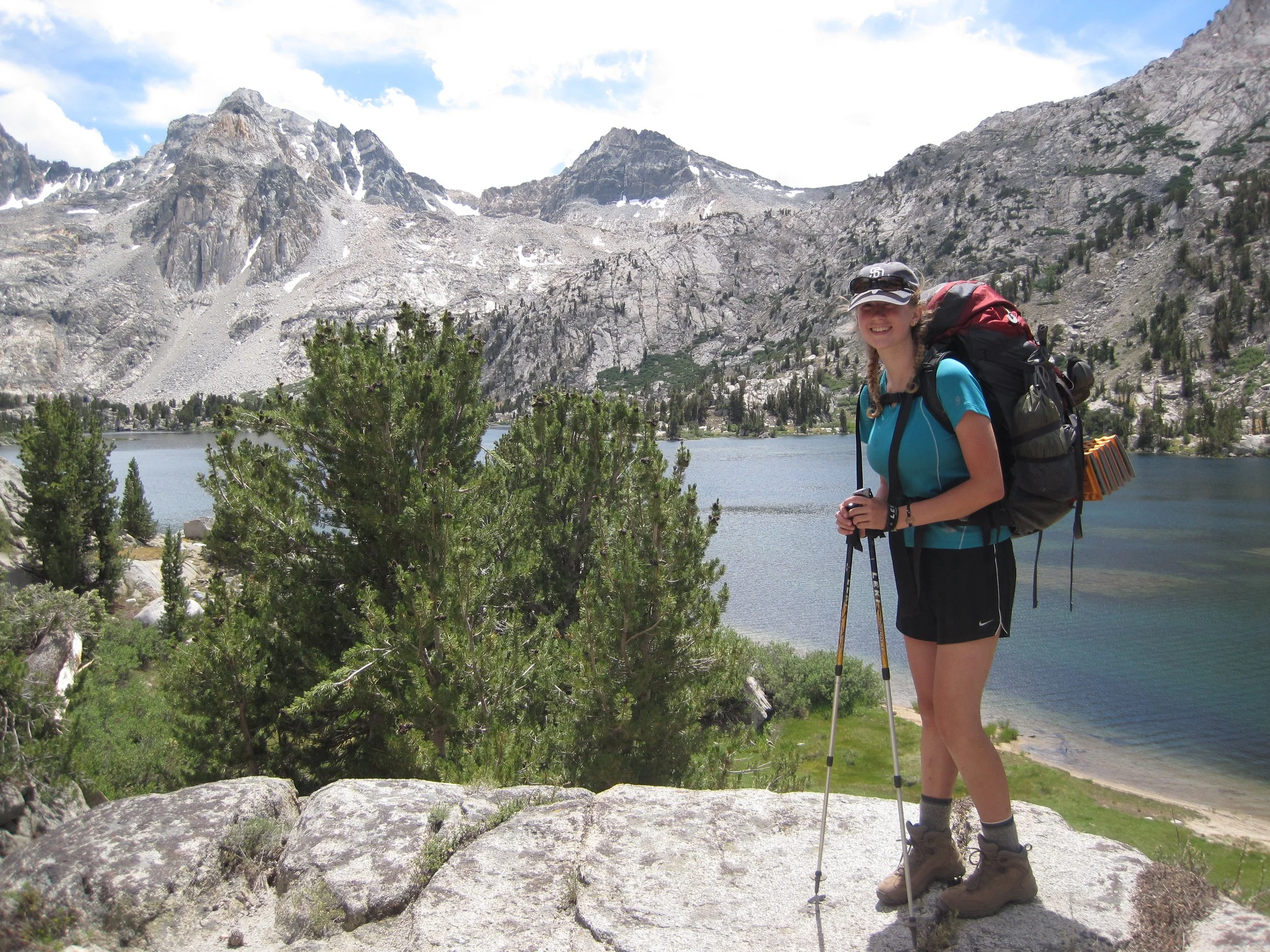 A Girl Scout backpacker on a rock ledge overlooking a turquoise lake with snow dotted mountains in the background.
