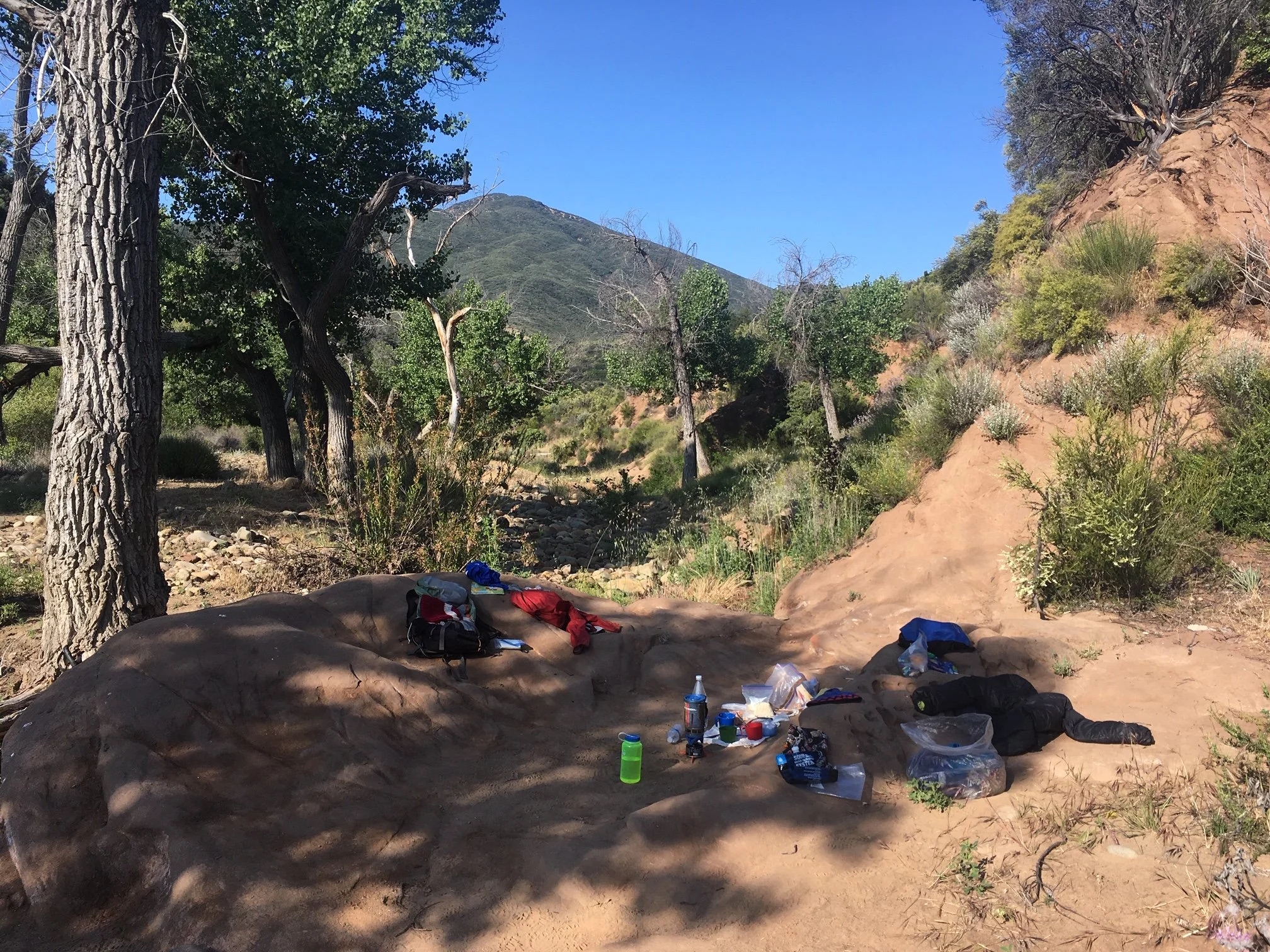 Backpacking equipment in a red dirt campsite surrounded by brush and a few trees.